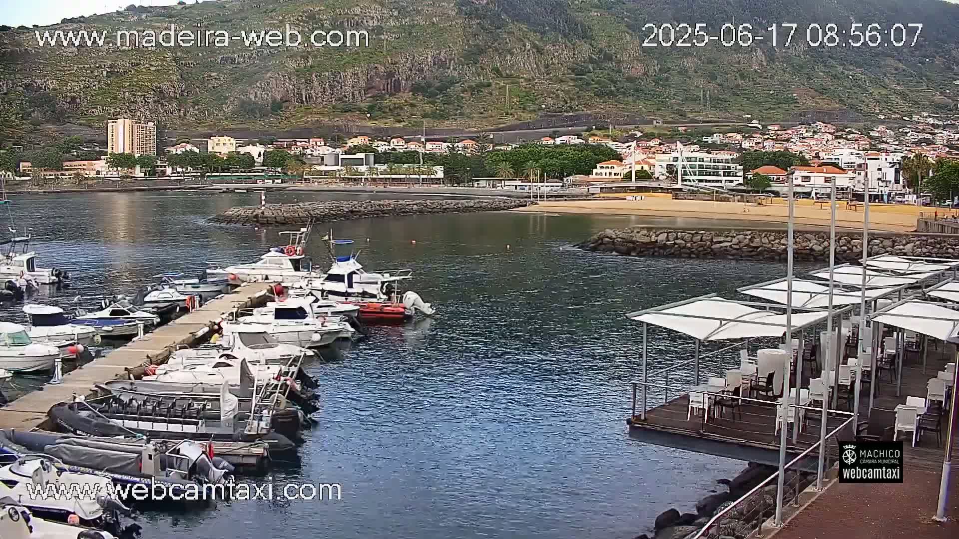 A marina filled with boats is situated next to a sandy beach and a town built on a hillside, under a mostly clear sky.
