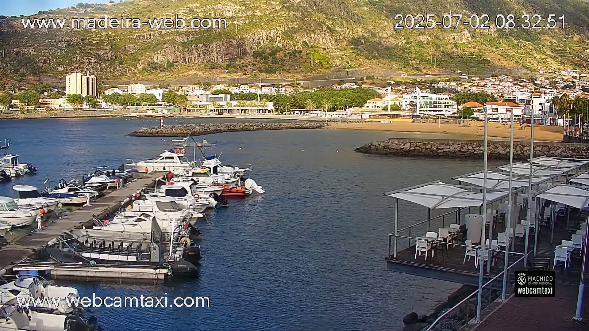A calm harbor filled with numerous boats is nestled beside a sandy beach and a town situated against a mountainside under a clear sky.