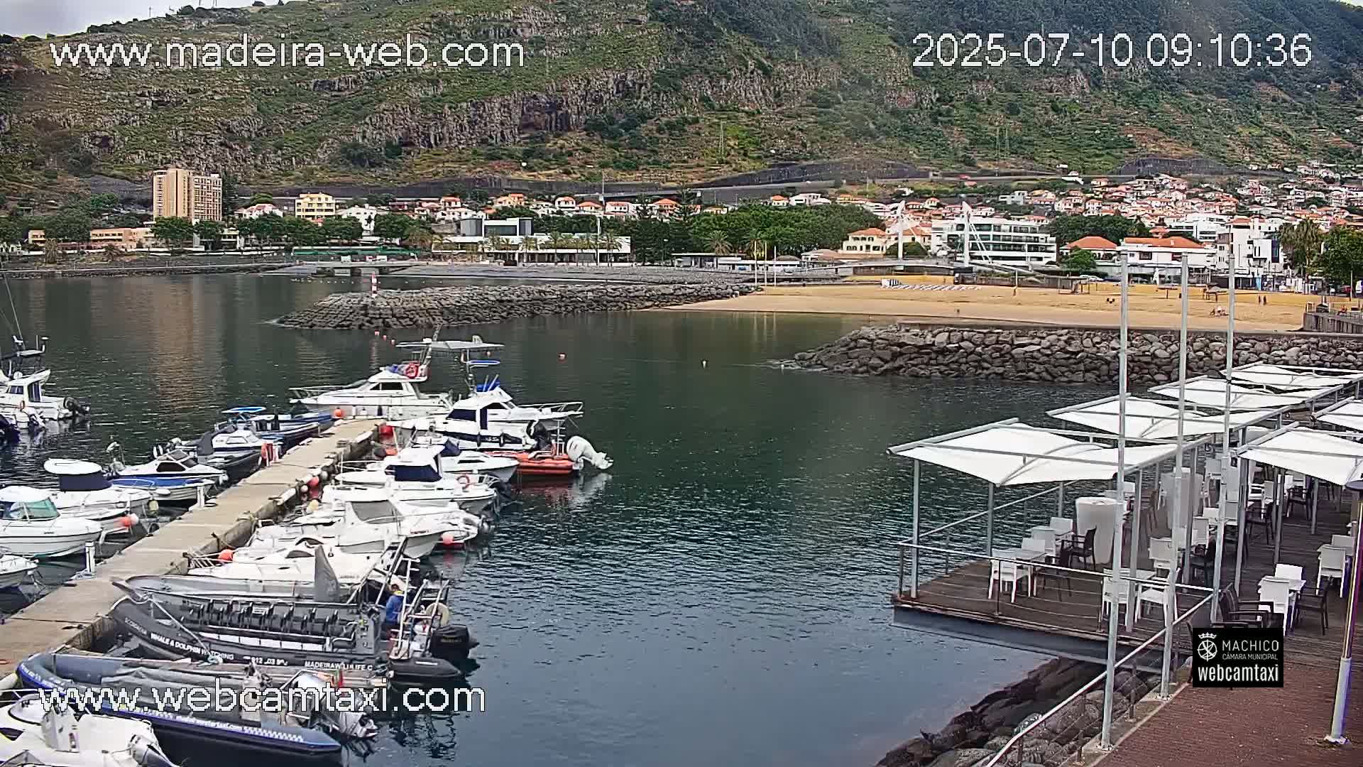 A harbor filled with numerous boats is situated next to a sandy beach and a small town nestled against a mountainous backdrop under overcast skies.