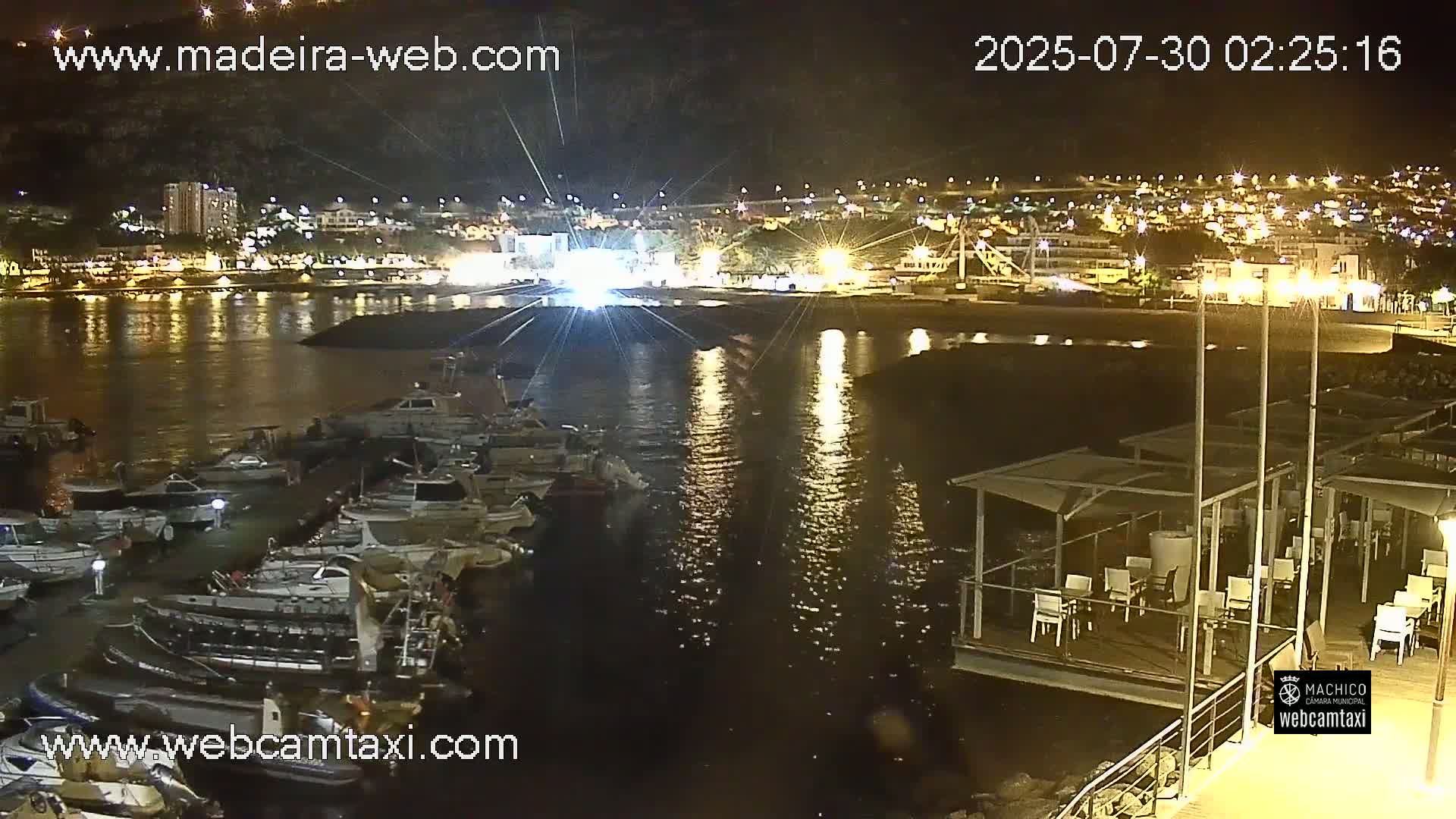A marina filled with numerous boats is situated in front of a town nestled against a hillside, under a mostly sunny sky.