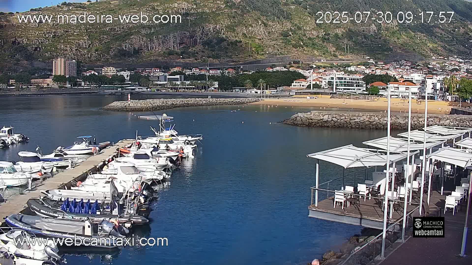 A marina filled with docked boats is situated near a sandy beach and a town built against a hillside on a sunny day.