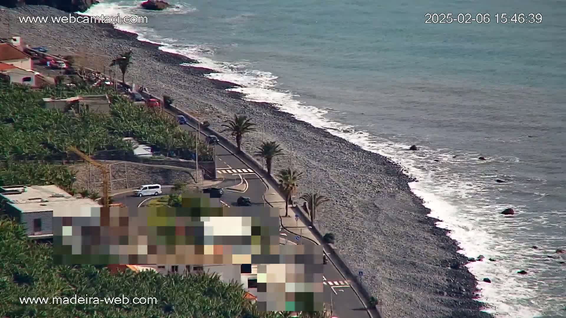 A rocky beach with gentle waves borders a coastal road lined with palm trees and buildings, partially obscured by a blurred section; banana plantations are visible inland.