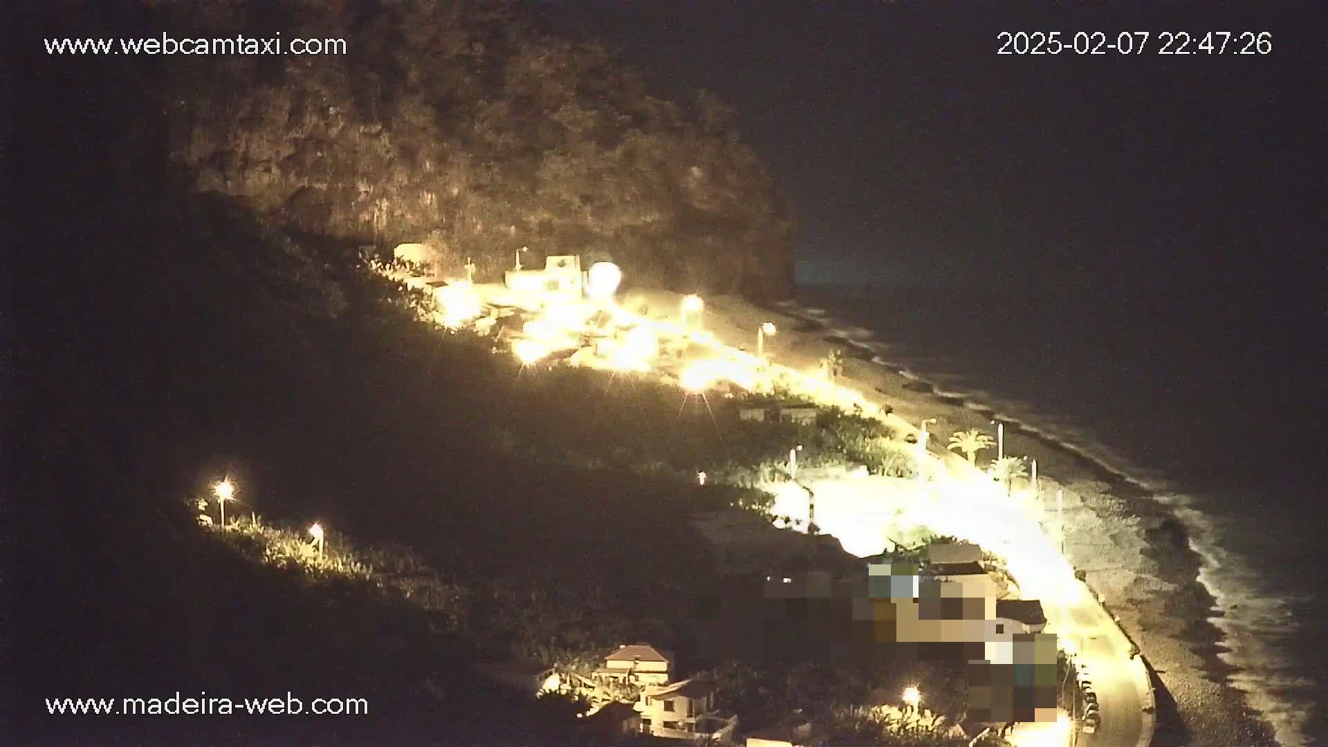 A nighttime coastal scene shows a dark hillside illuminated by lights near a pebble beach with gentle waves under a clear, dark sky.