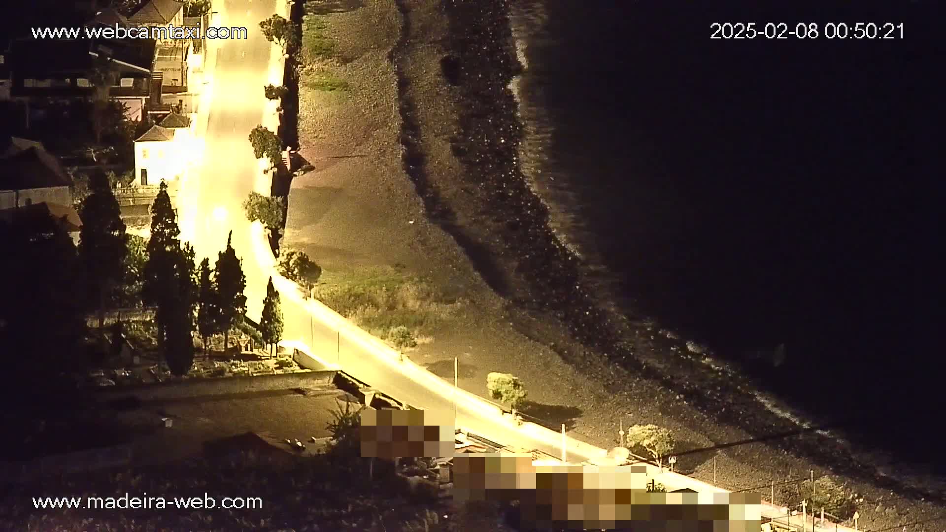 A nighttime, high-angle view shows a dark, rocky coastline with gentle waves lapping the shore, next to a brightly lit road winding through a town with dark green trees.