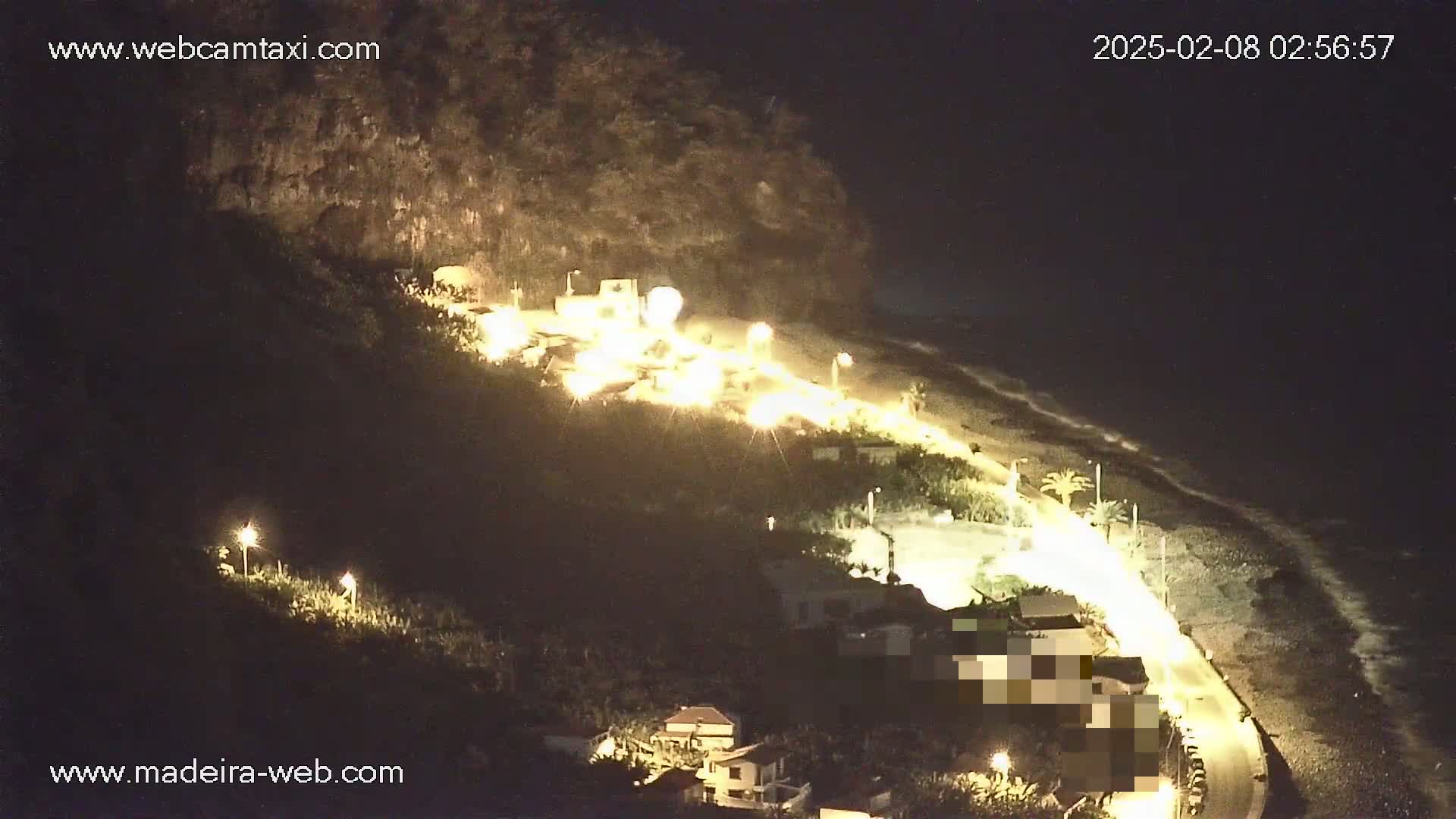 A nighttime coastal scene shows a road winding along a beach below a hillside with illuminated buildings and vegetation.