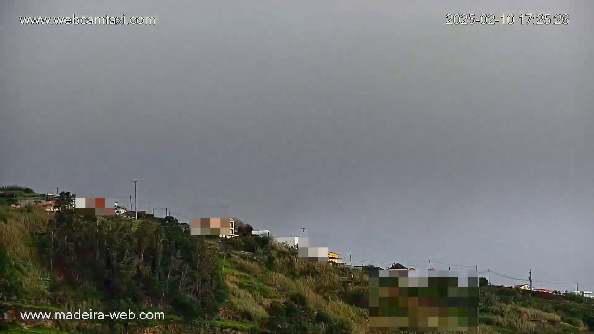 Under an overcast sky, a hillside covered in vegetation displays several buildings, with some portions obscured.