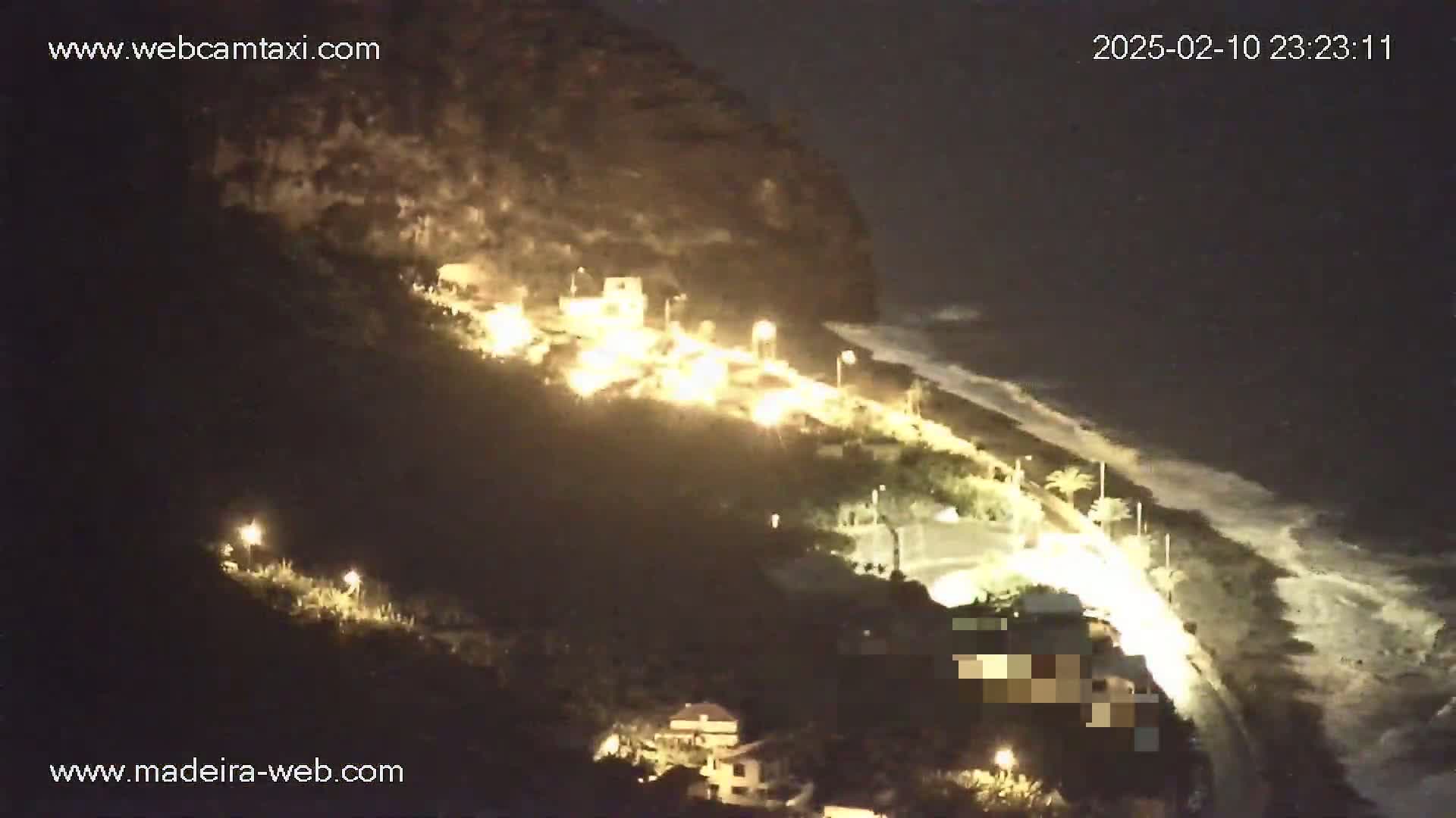A nighttime coastal scene shows a dark, rocky hillside with illuminated buildings and a road winding along a beach with waves under a dark sky.