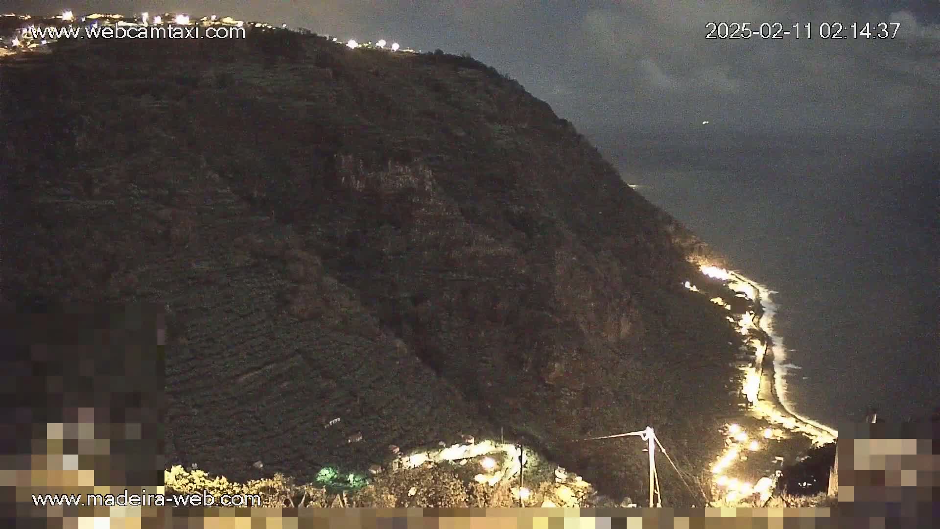 A nighttime view of a dark, verdant cliffside overlooking a coastline with a road illuminated by lights under a partly cloudy sky.