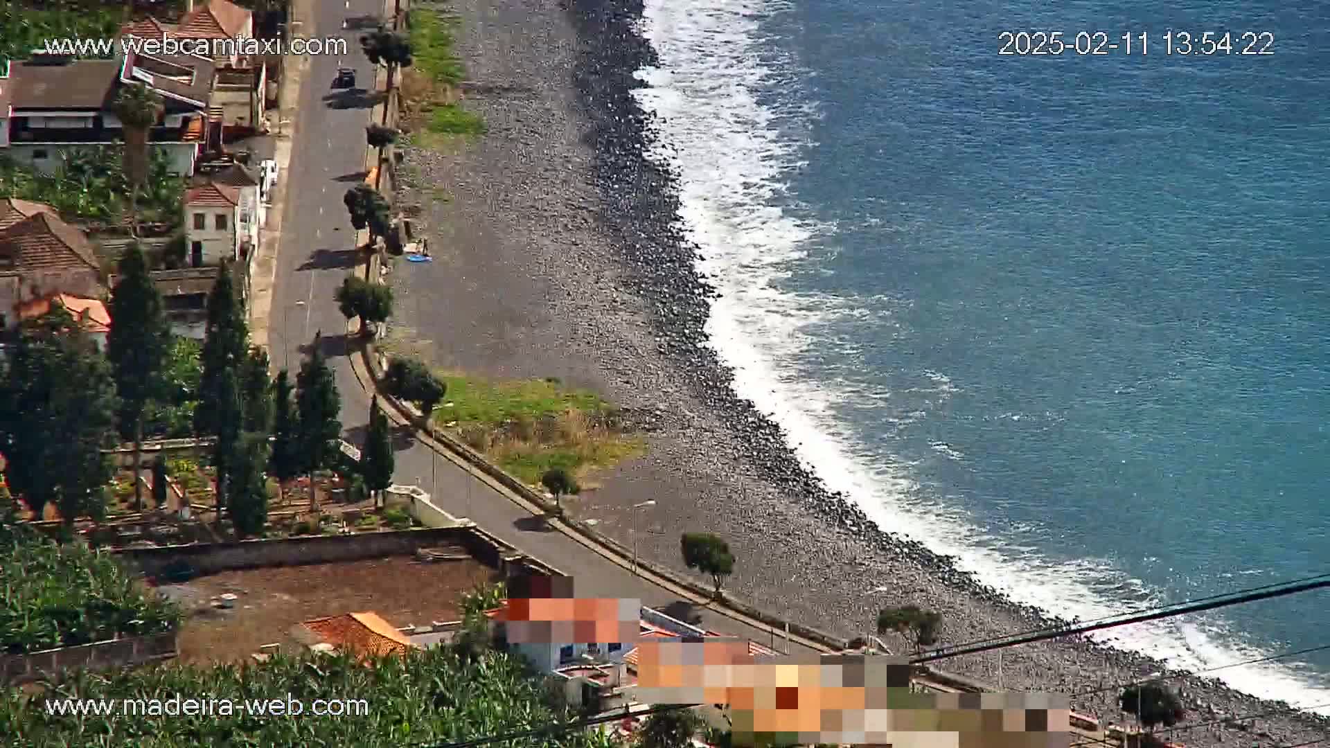 An aerial view shows a dark-colored, rocky beach with small waves lapping the shore next to a road lined with houses and trees on a sunny day.