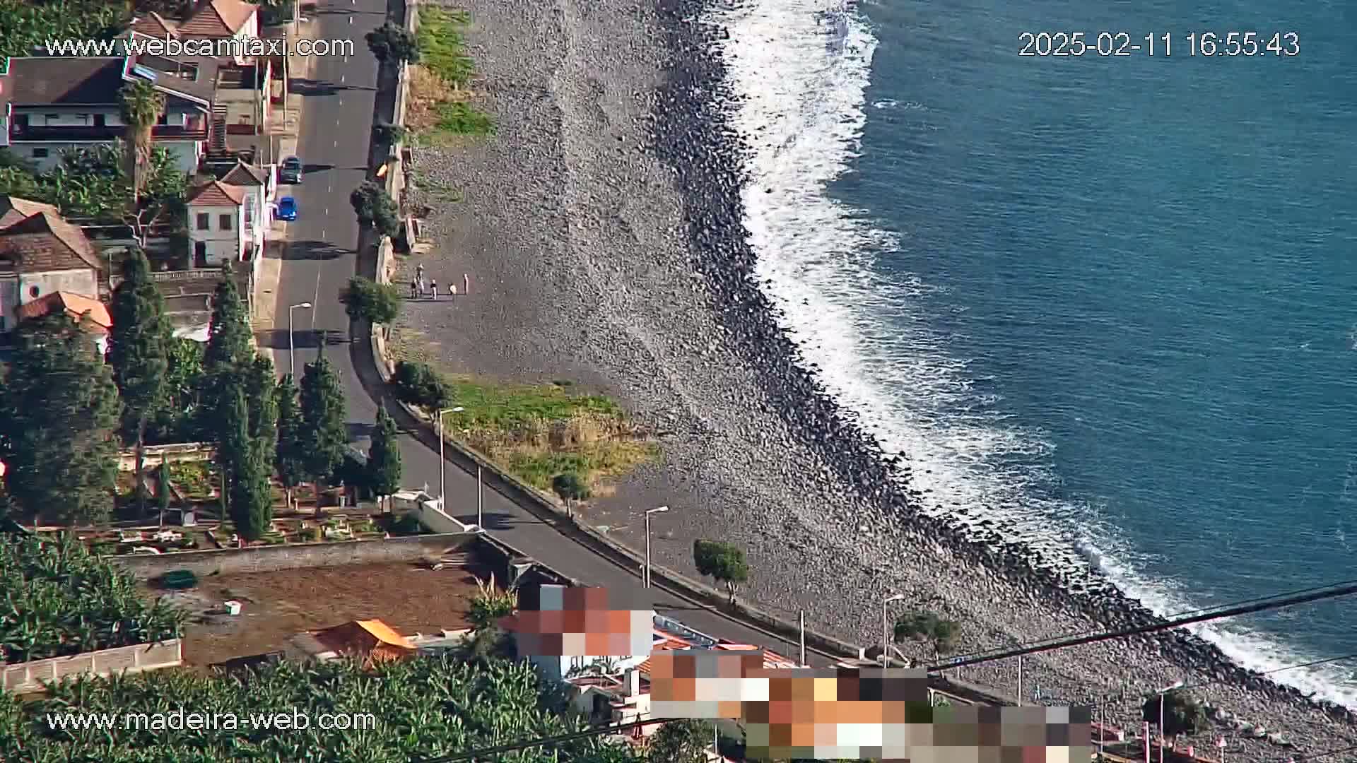 A rocky beach with small waves meets a road winding through a residential area with a cemetery and vegetation.