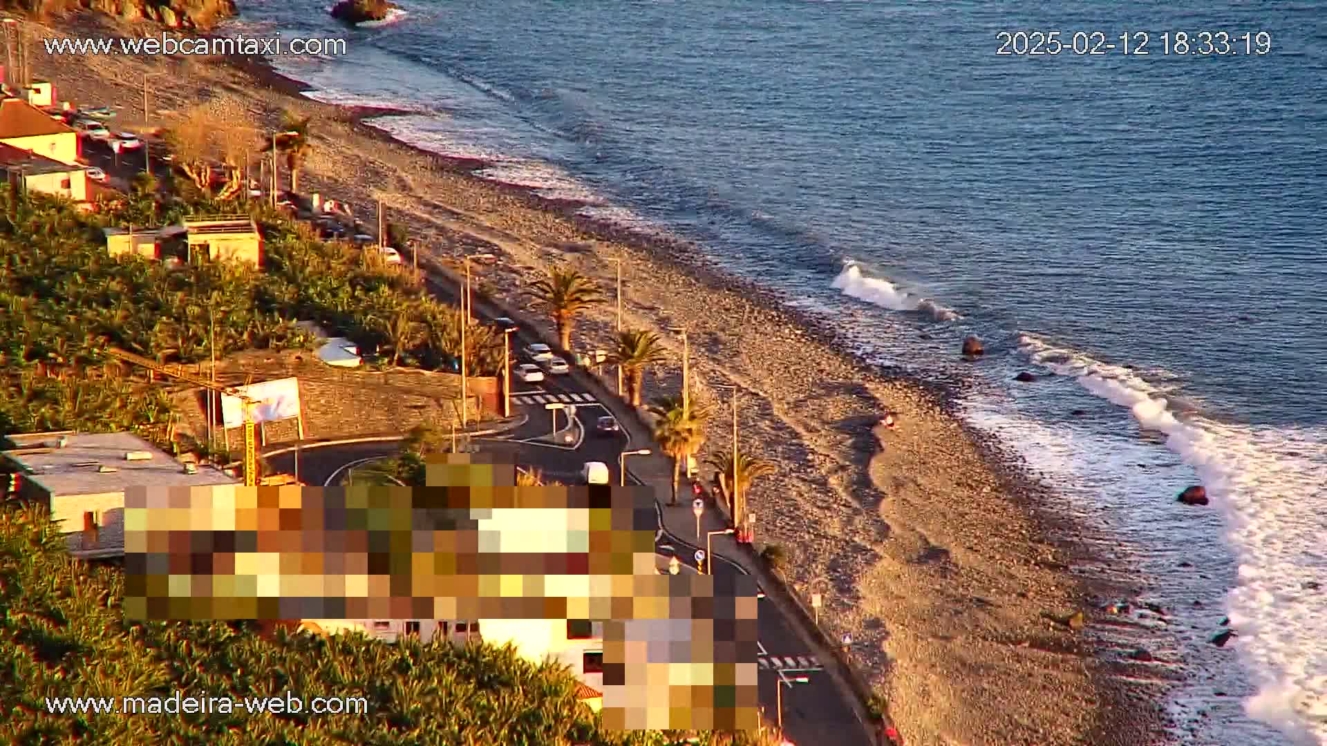 An aerial view shows a pebble beach with gentle waves lapping the shore, next to a road lined with palm trees and buildings.