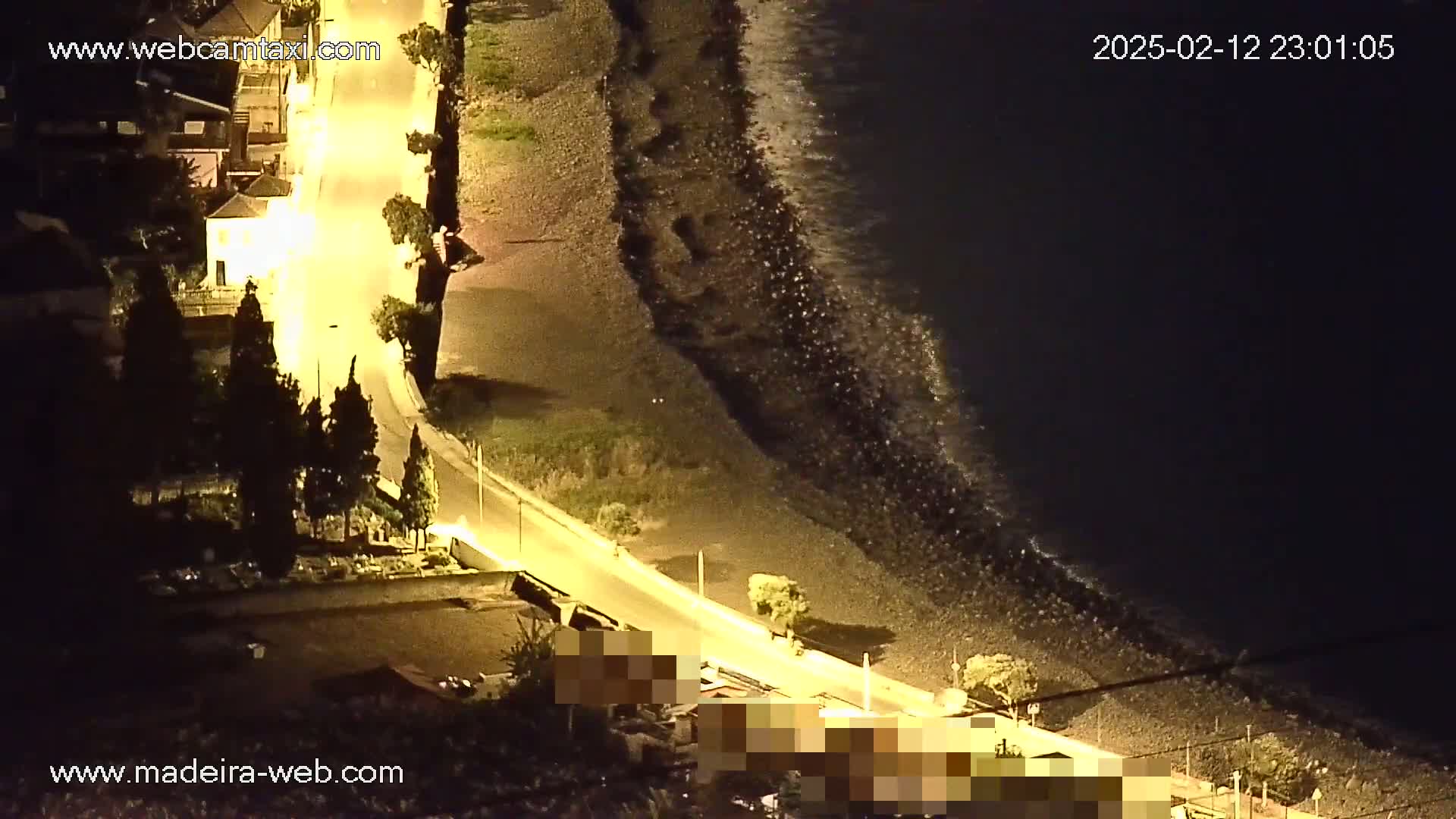 An illuminated road curves down to a dark, rocky beach at night, where small waves lap the shore under a dark sky.