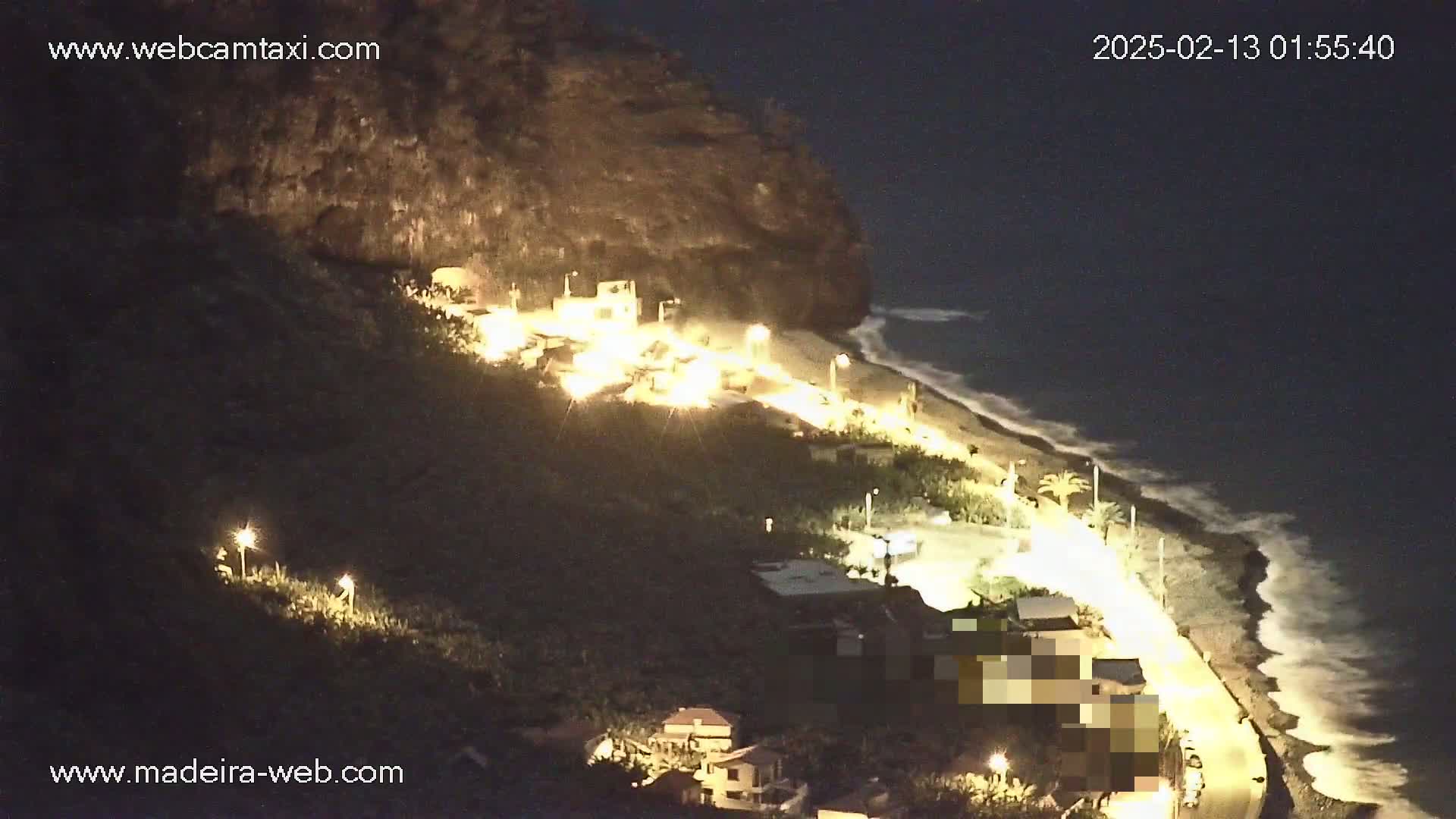 A nighttime coastal scene shows a road winding along a beach with gentle waves under a dark sky, illuminated by streetlights and building lights on the hillside above.