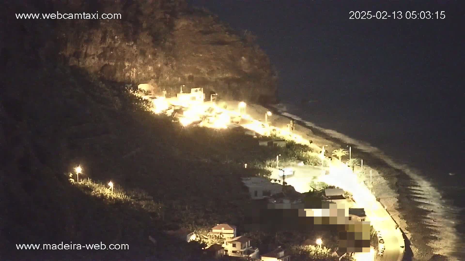 A nighttime, coastal scene shows a dark, rocky cliff overlooking a beach with a road and buildings illuminated by artificial lights.