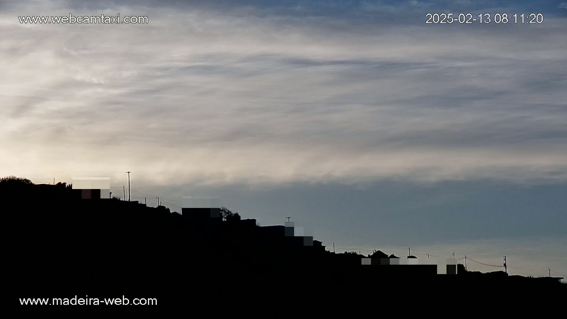 Under a mostly cloudy sky, a hillside silhouetted against the horizon shows a line of dark buildings.