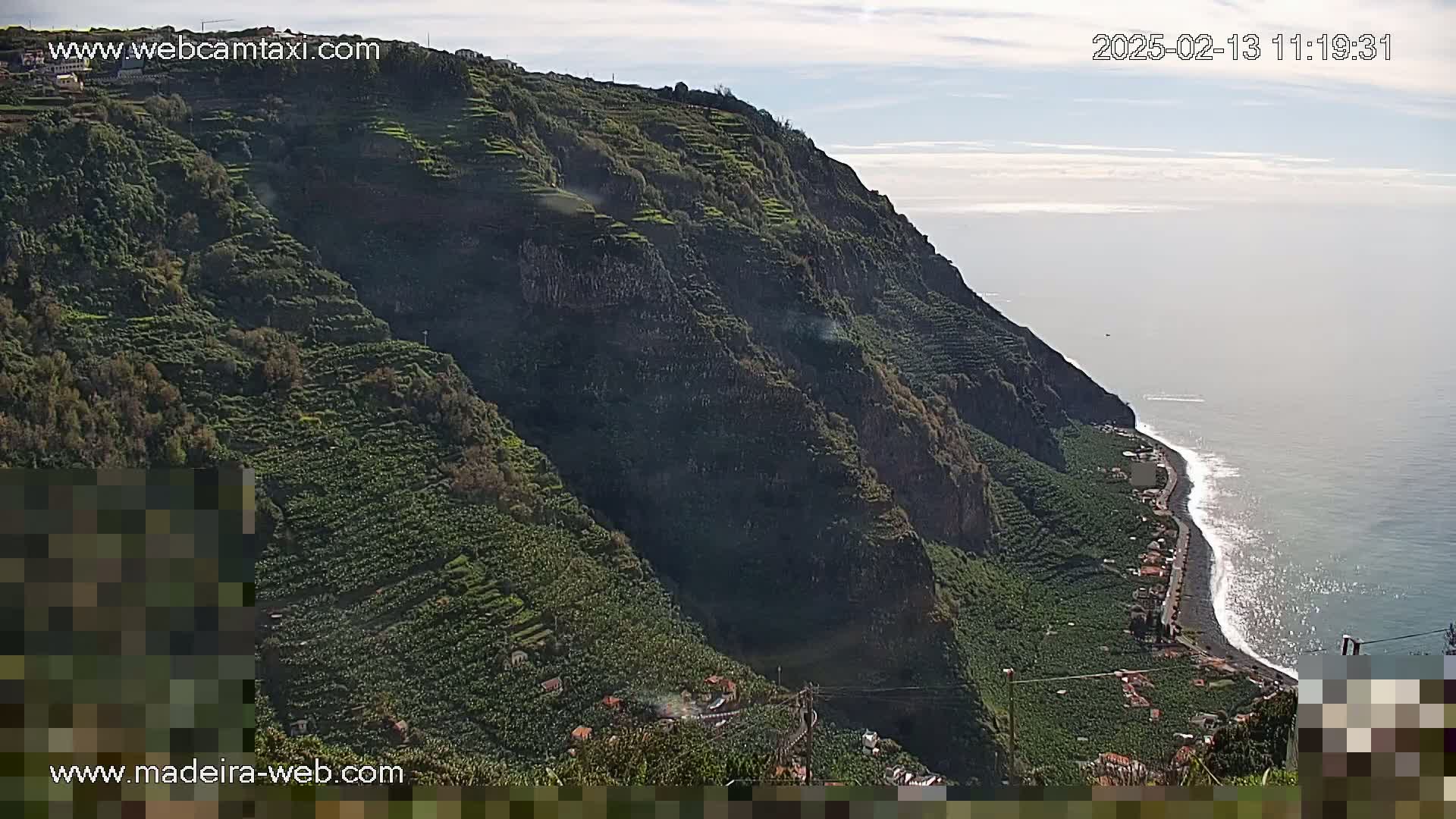 A steep, verdant hillside terraced with crops descends to a dark-sand beach and ocean under a mostly clear sky.