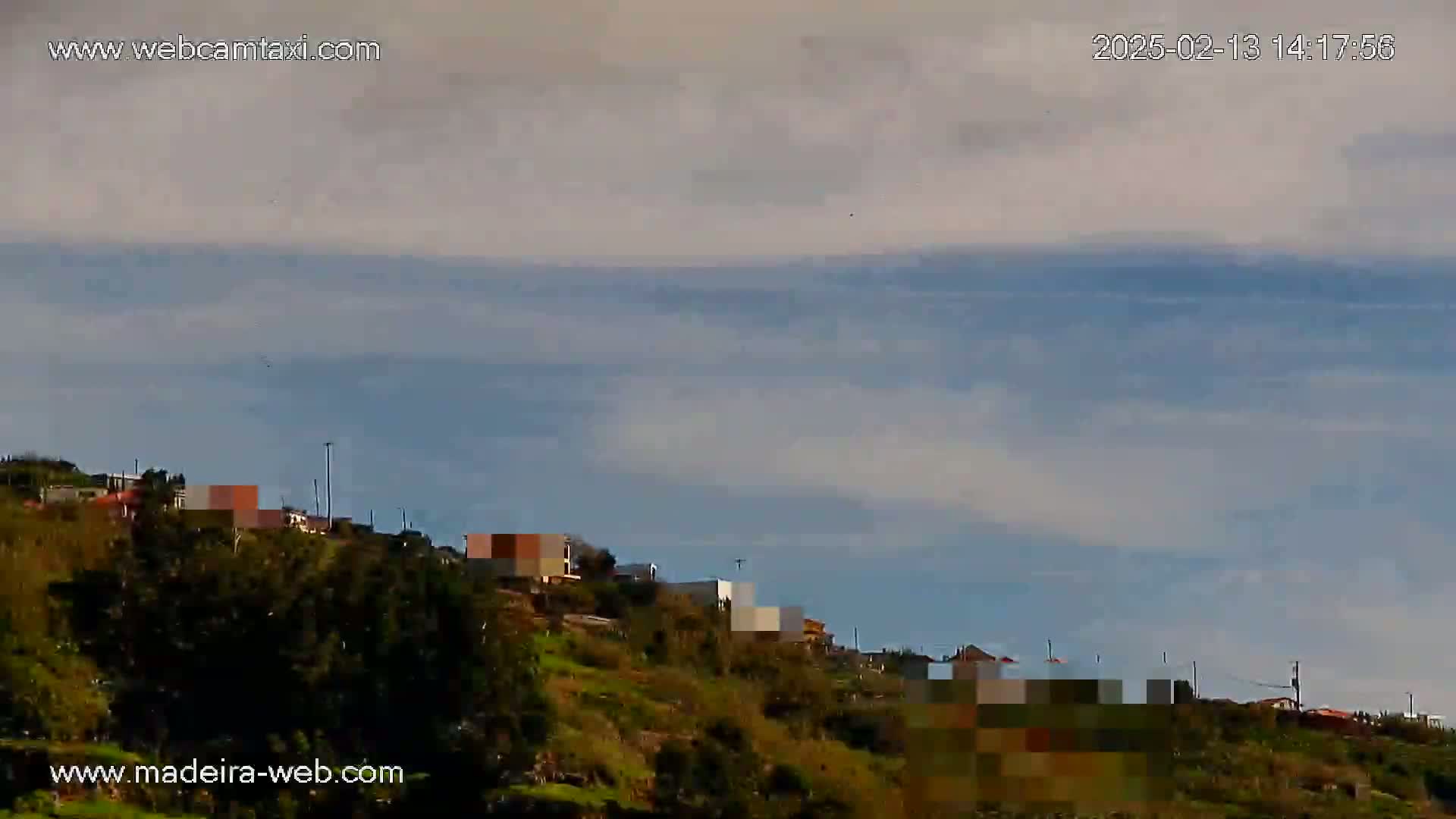 A hillside covered in green vegetation with a few buildings visible under a mostly cloudy sky.