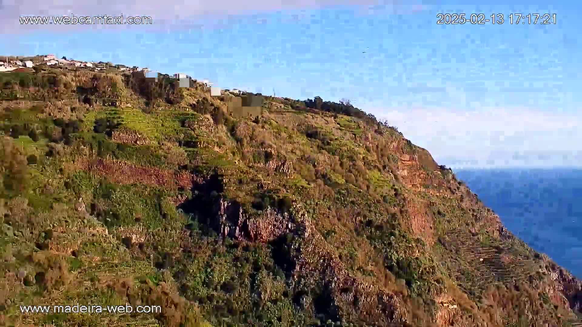 A steep, terraced hillside with sparse vegetation and a small village at the top overlooks a calm ocean under a clear blue sky.