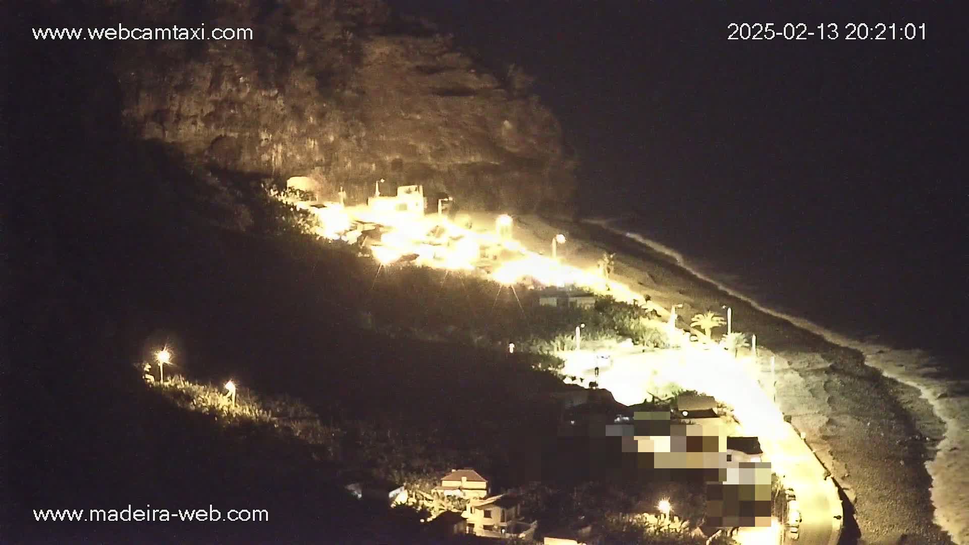 A nighttime, coastal scene shows a dark, rocky hillside illuminated by artificial lights, with a dimly lit beach and ocean visible below.