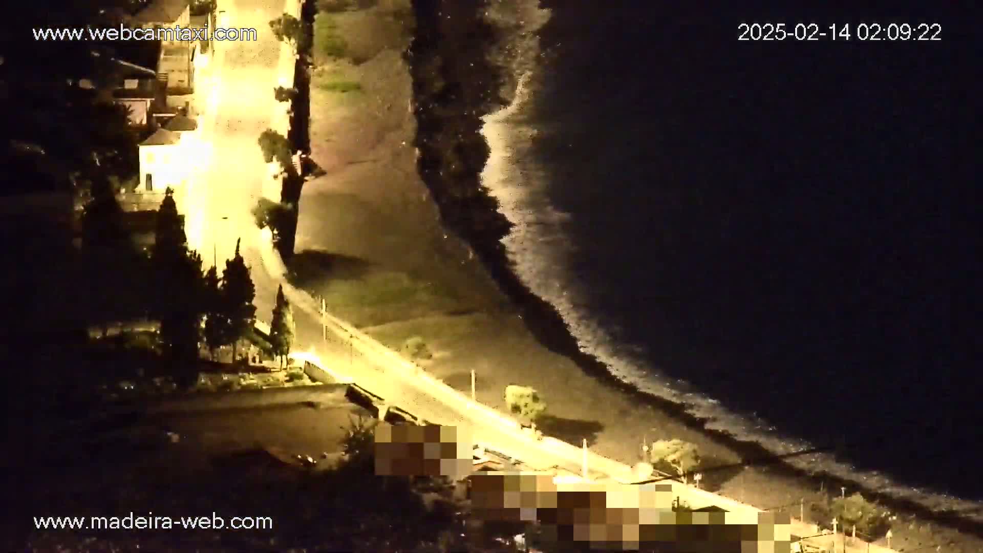 A nighttime, high-angle view shows a dark ocean with waves lapping a shoreline next to a brightly lit road and buildings.