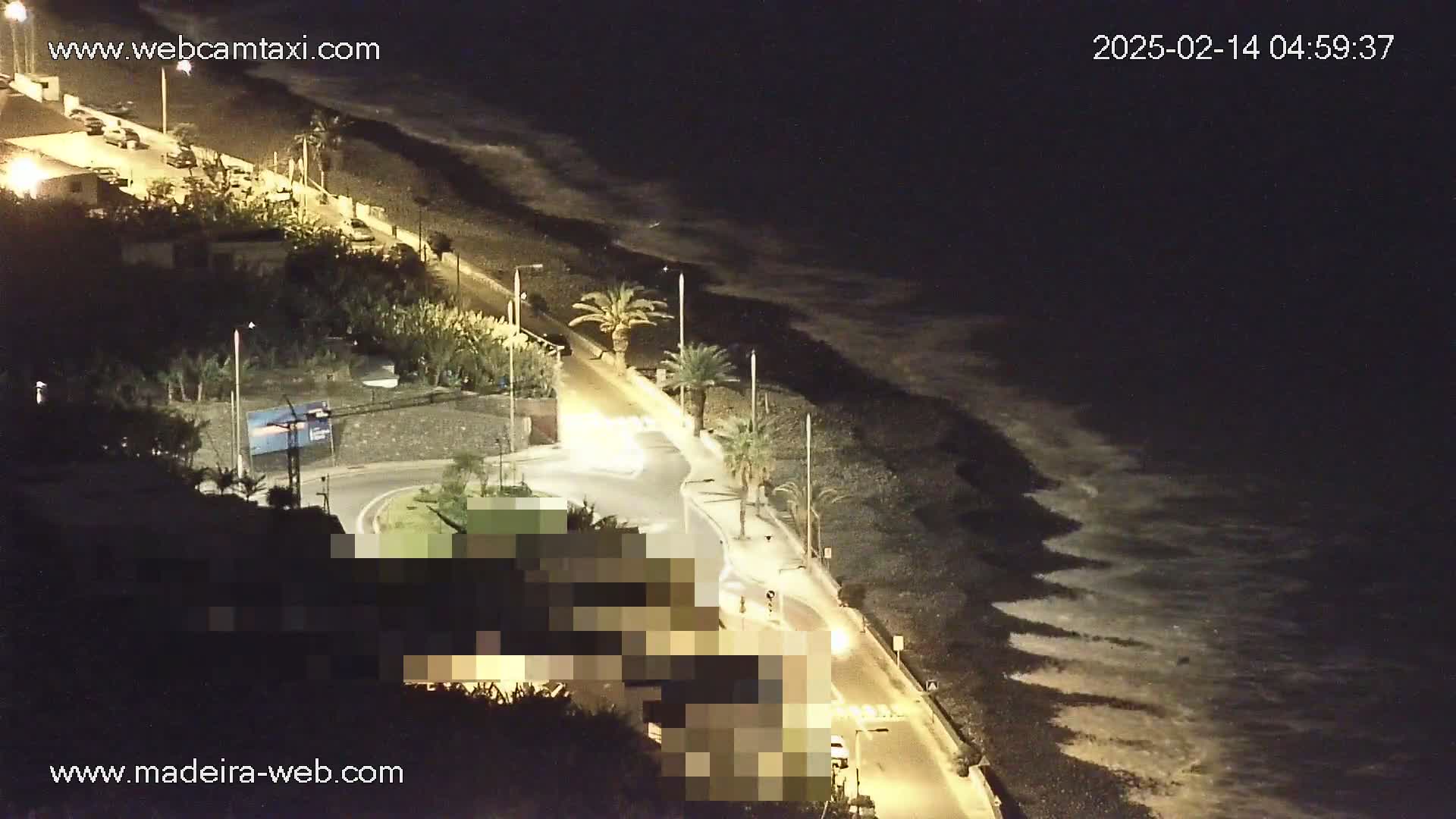 A nighttime coastal road scene shows gently lapping waves on a dark, pebble beach next to a road with parked cars and palm trees under artificial light.