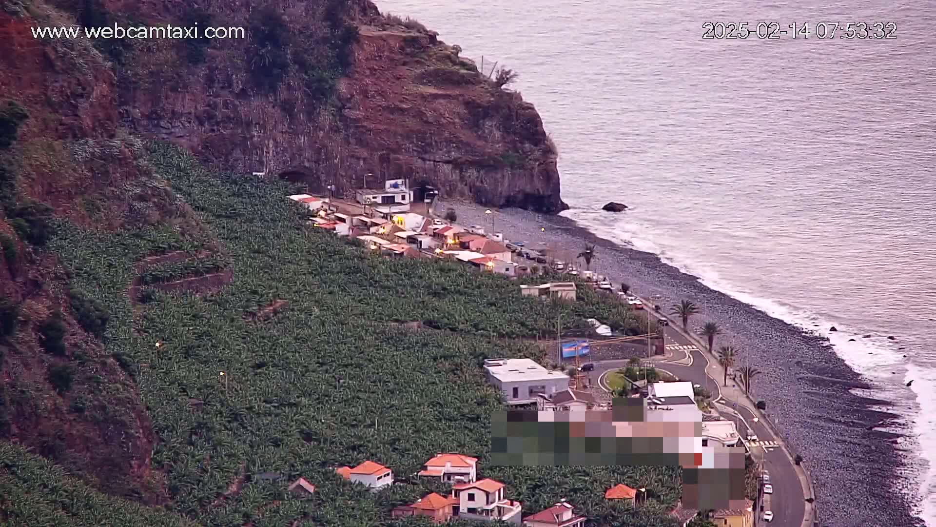 A coastal village nestled beside a dark-sand beach and banana plantations, situated beneath a steep cliff face on a calm, overcast day.