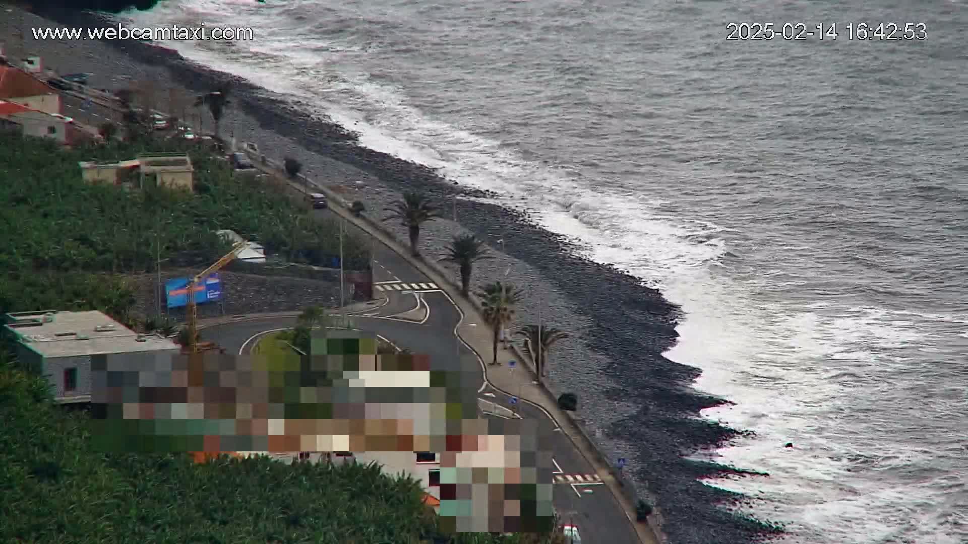 A dark-colored, rocky beach with moderate waves laps against the shore next to a coastal road lined with palm trees and buildings.