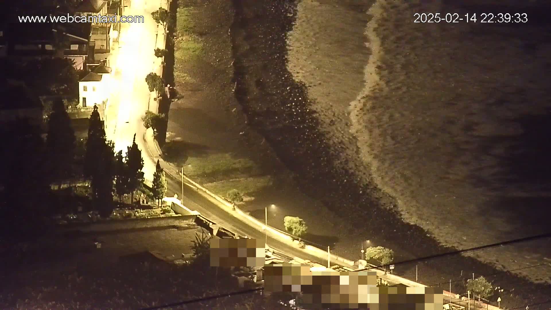 Large waves crash against a shoreline at night, next to a road and buildings illuminated by streetlights.