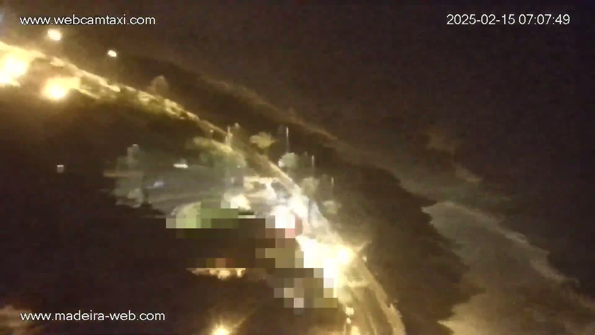 An aerial nighttime view shows a road with vehicles and surrounding vegetation, illuminated by streetlights and vehicle headlights under a dark, cloudy sky.