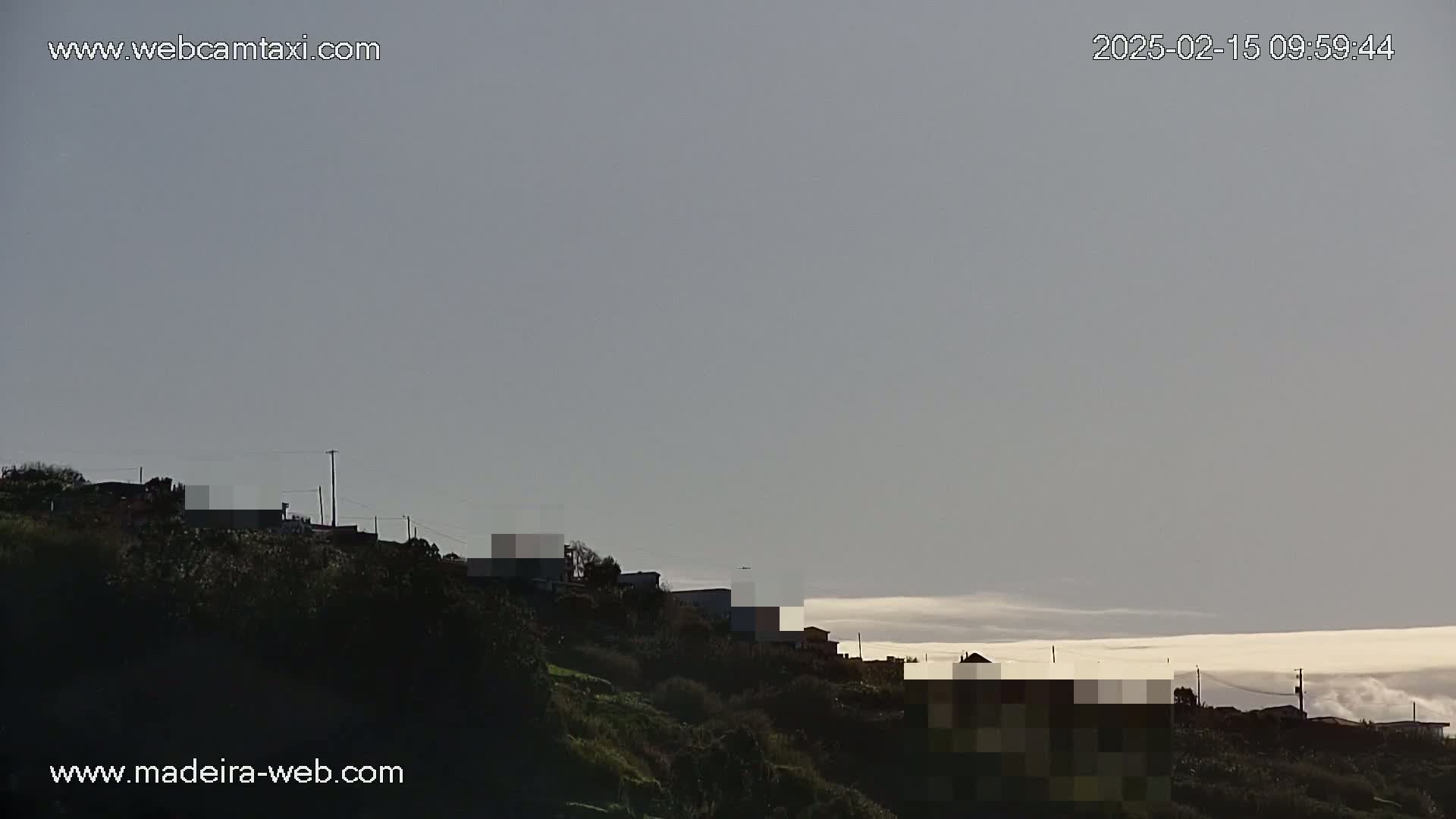 A hillside covered in dark green vegetation shows a few buildings, partially obscured by a low-lying layer of clouds under a pale, overcast sky.
