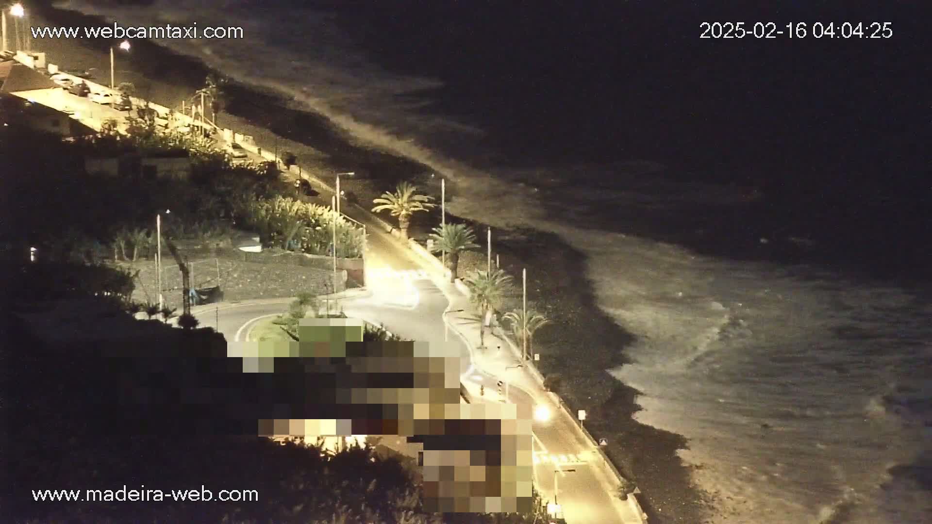 A nighttime coastal scene shows a road curving alongside a dark beach with waves lapping the shore under a dark sky.