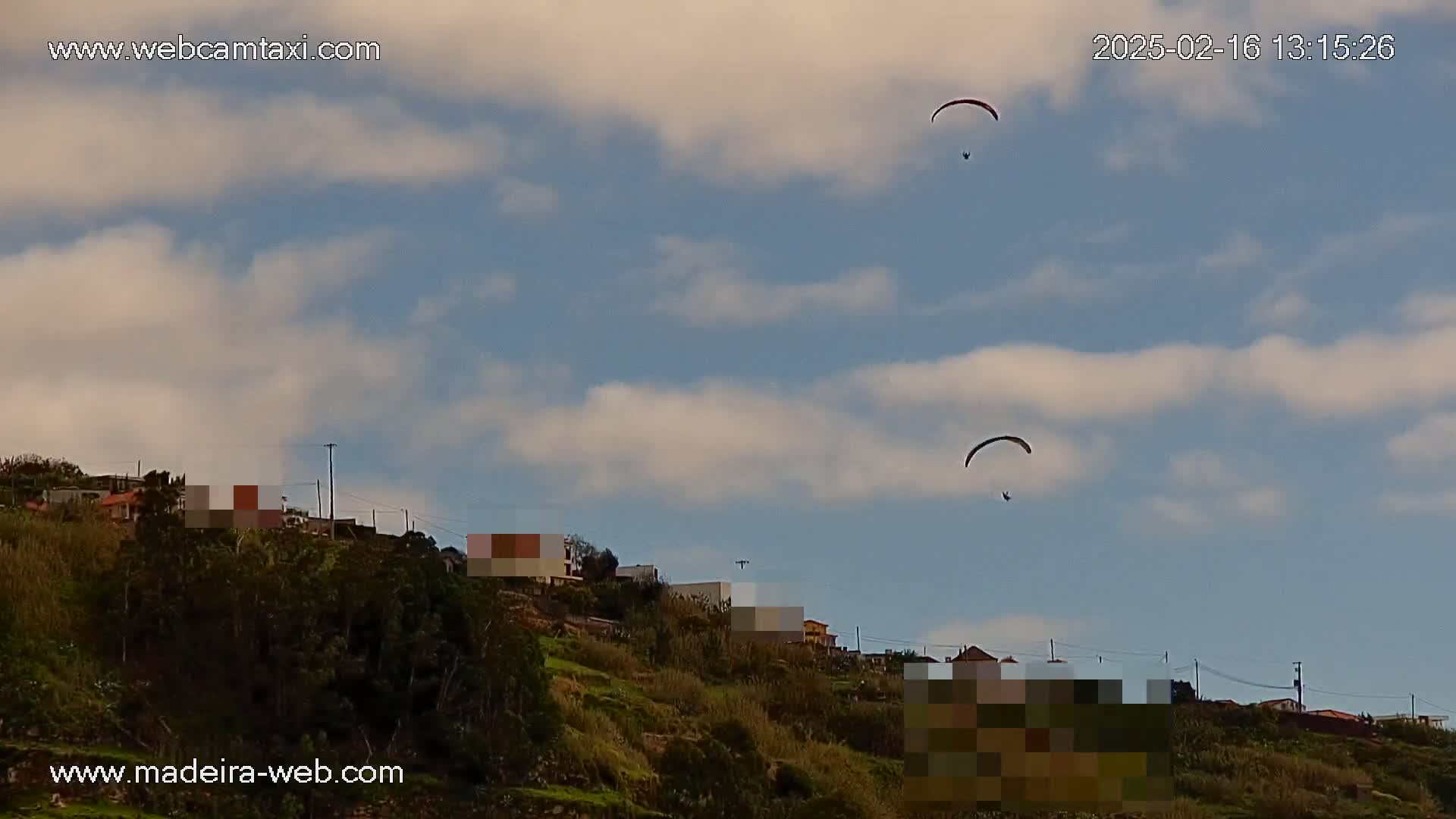 Two paragliders fly in a partly cloudy sky above a hillside with buildings and vegetation.