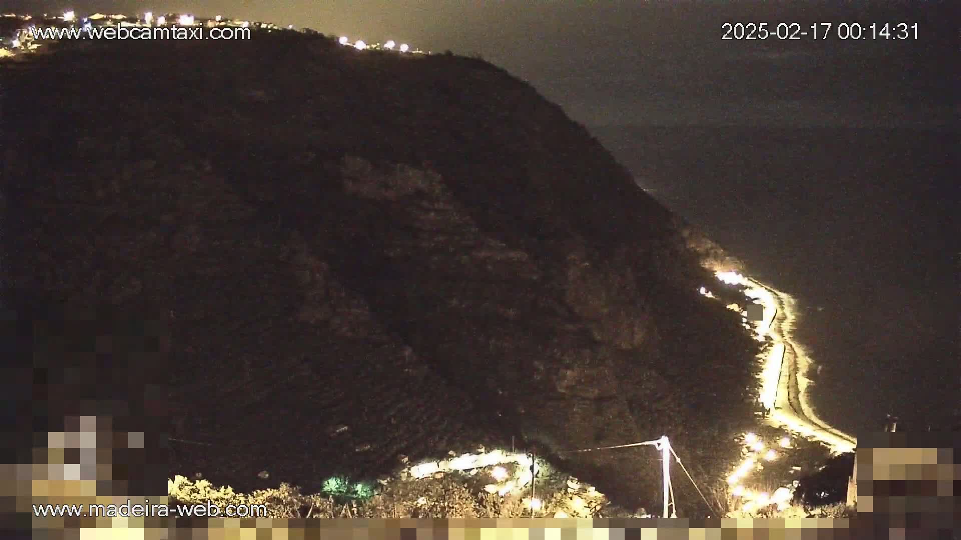 A nighttime view of a dark, mountainous coastline with a winding road illuminated by vehicle headlights, overlooking a dark body of water.