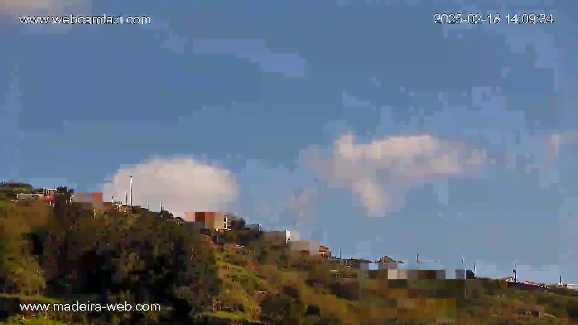 A mostly clear blue sky with a few fluffy white clouds above a hillside village with green vegetation.