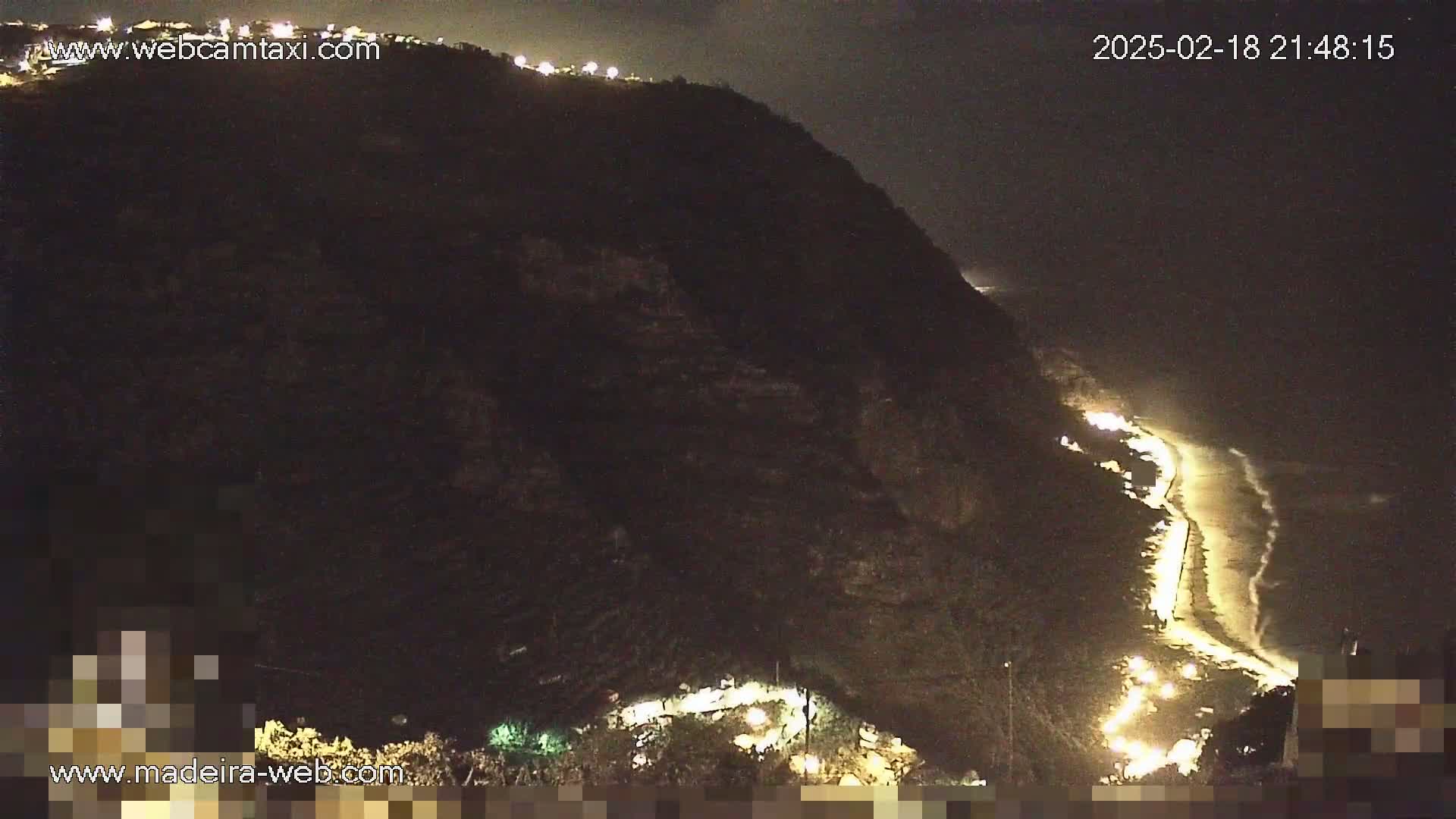 A nighttime view of a dark, mountainous coastline with a lit road winding down to a beach where waves are gently lapping the shore.