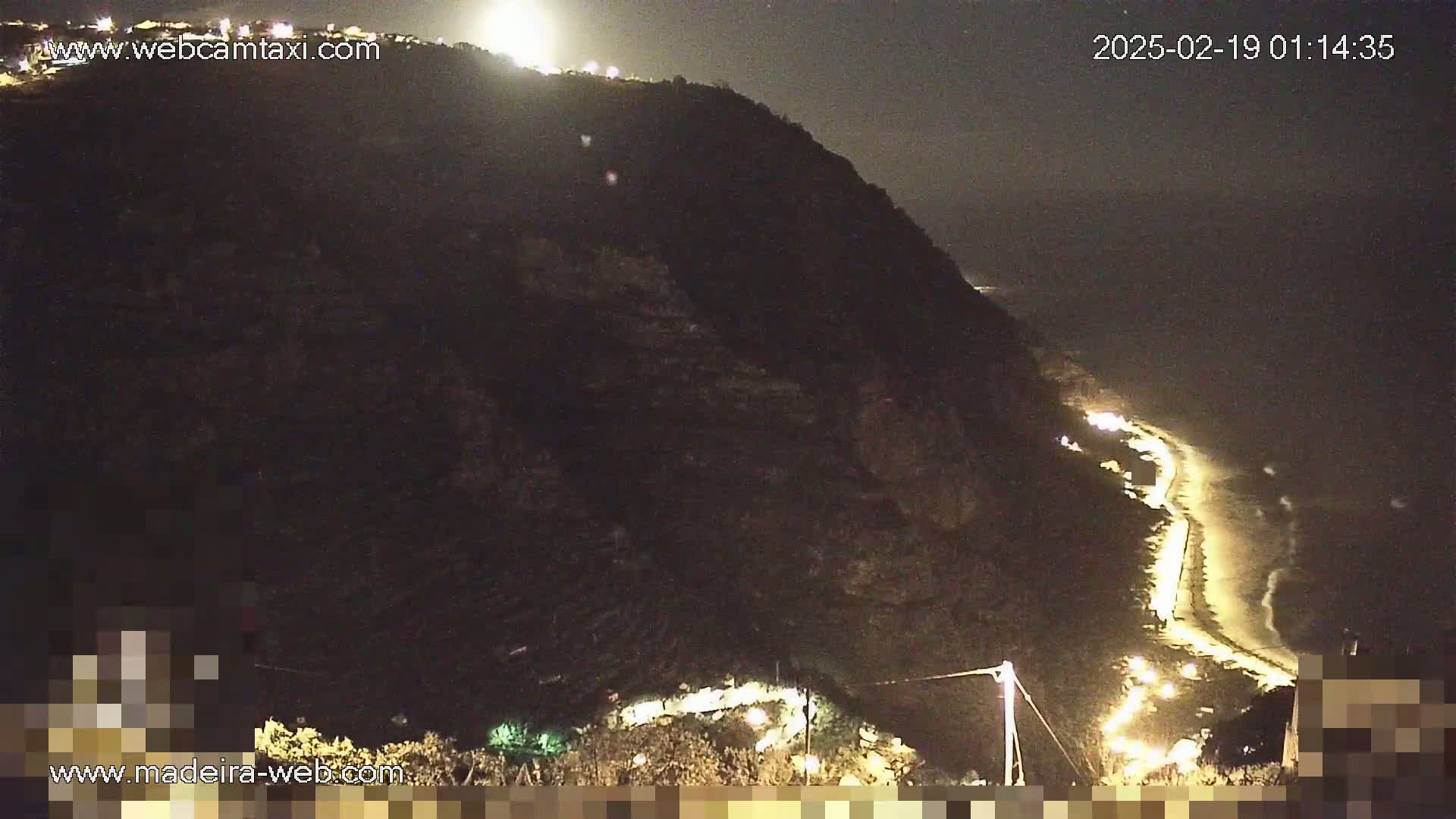 A nighttime view of a dark, cliffside coast with a road lit by lights winding along the beach.