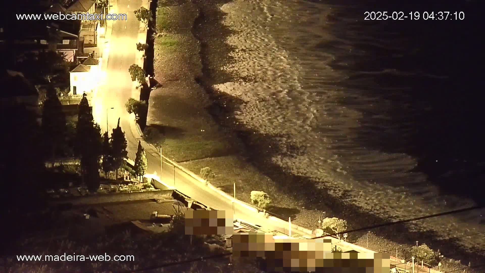An illuminated coastal road at night curves alongside a dark, rocky beach with moderately sized waves crashing on the shore.