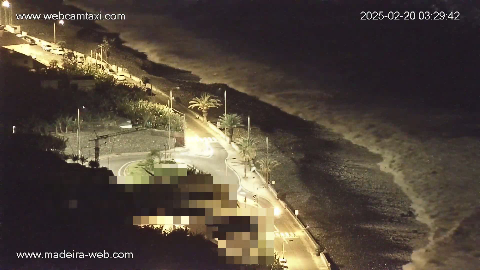 A nighttime, overhead view shows a coastal road with parked cars, palm trees, and a dark ocean with waves crashing on a pebble shore.