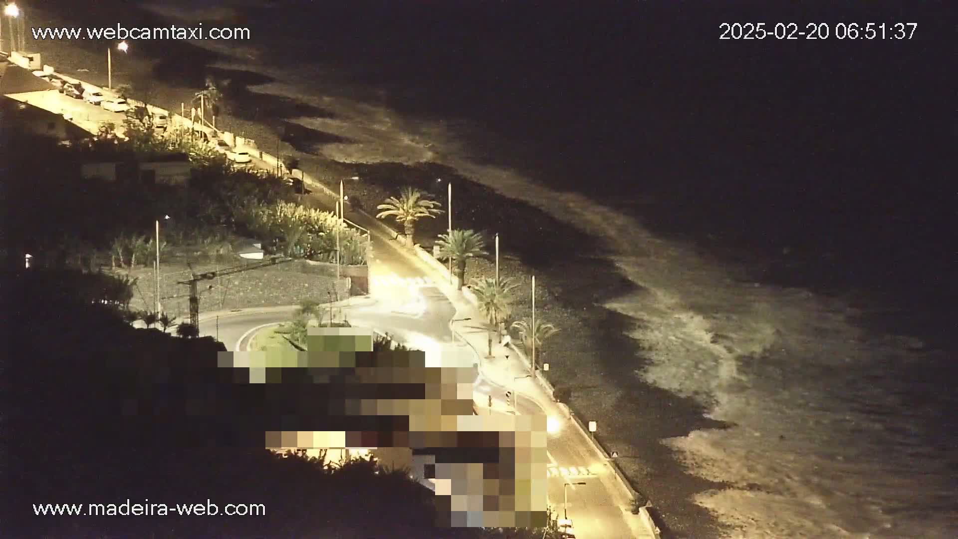 A nighttime coastal scene shows a road next to a dark beach with waves gently lapping the shore, lit by streetlights.