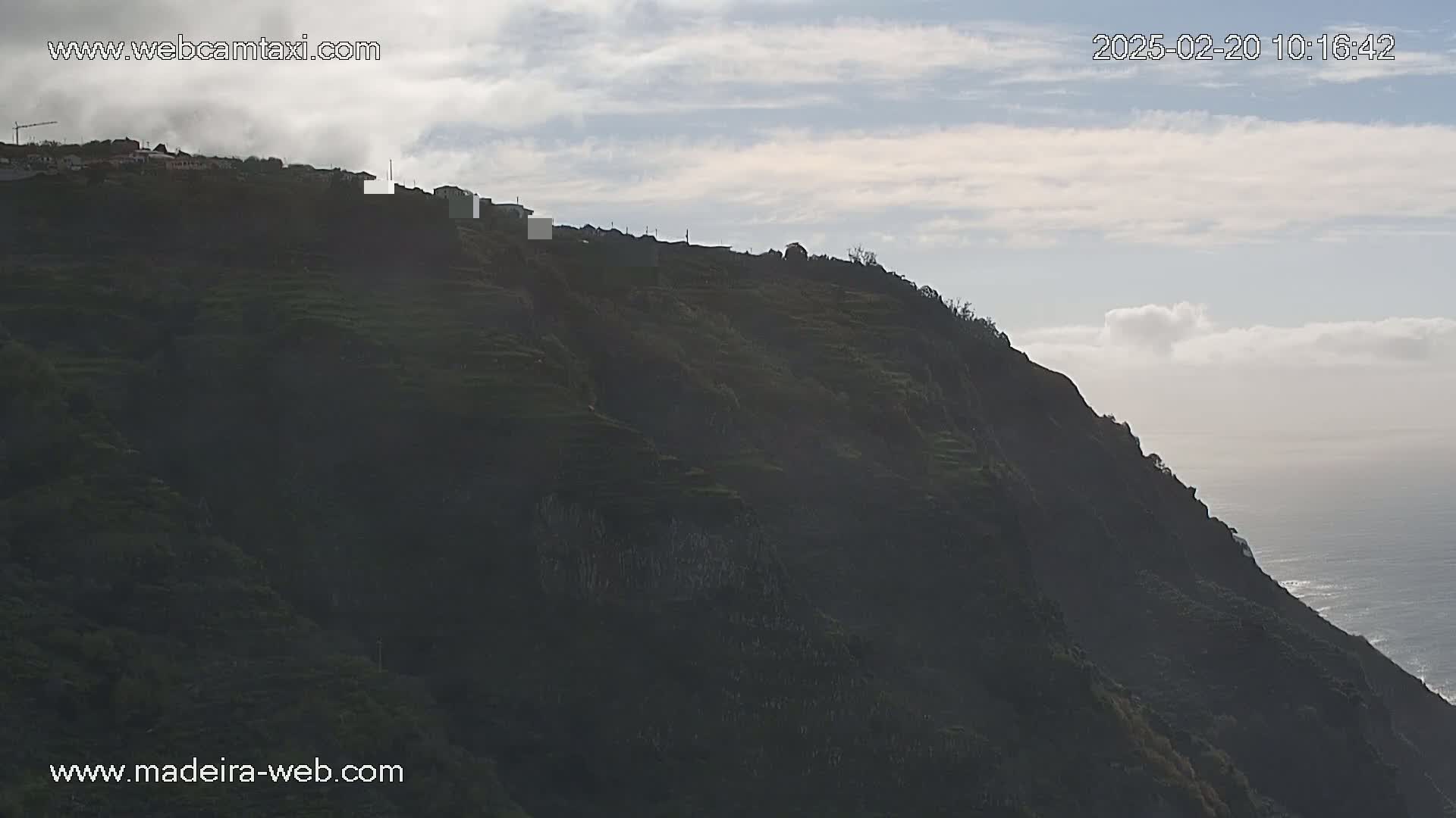 A dark, terraced hillside overlooks a calm ocean under a mostly cloudy sky.