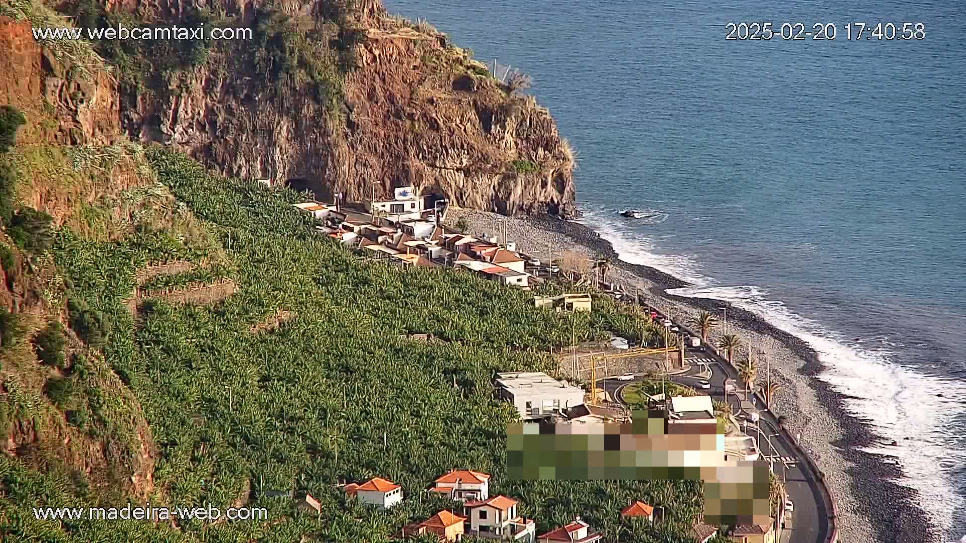 A coastal village nestled among banana plantations on a rocky hillside overlooking a dark-sand beach and calm ocean.