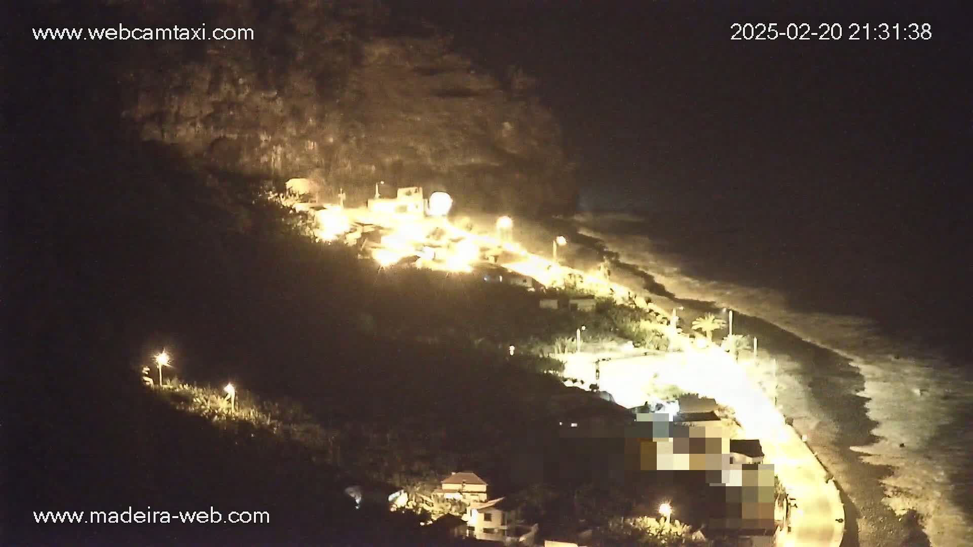 A nighttime, overhead view of a coastline with buildings and lights illuminating a dark beach and rolling ocean waves.