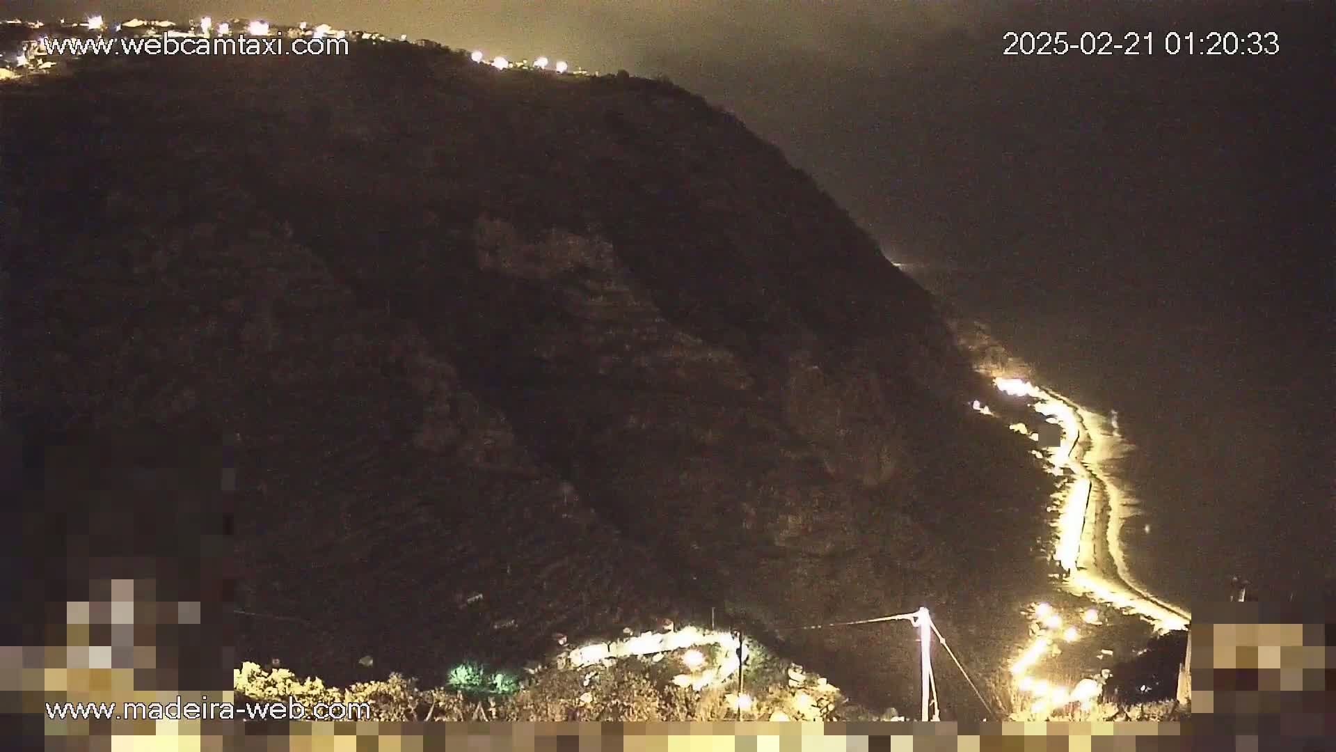 A nighttime view of a dark hillside illuminated by lights along a winding road that leads to a dimly lit beach.