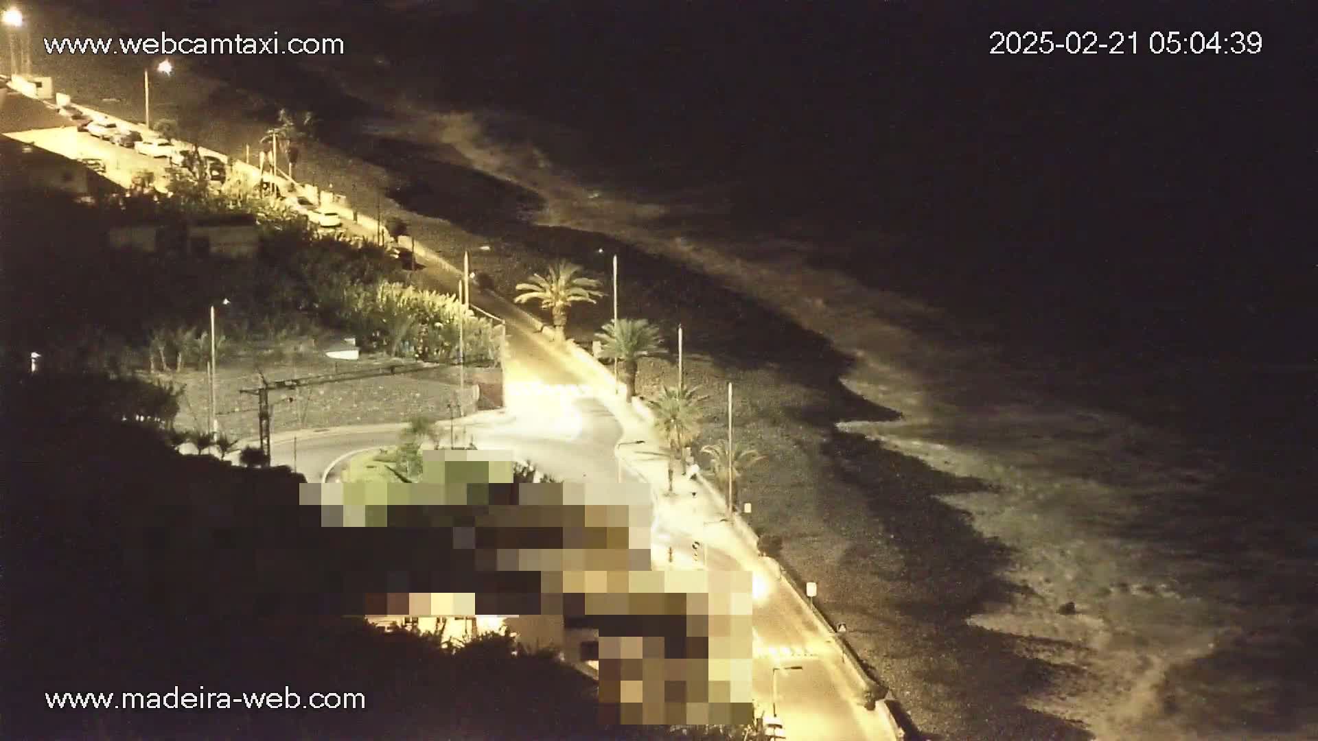 A nighttime, high-angle view shows a dark, pebble beach with gentle waves lapping the shore, a road lined with palm trees and buildings, and several vehicles parked along the roadside.