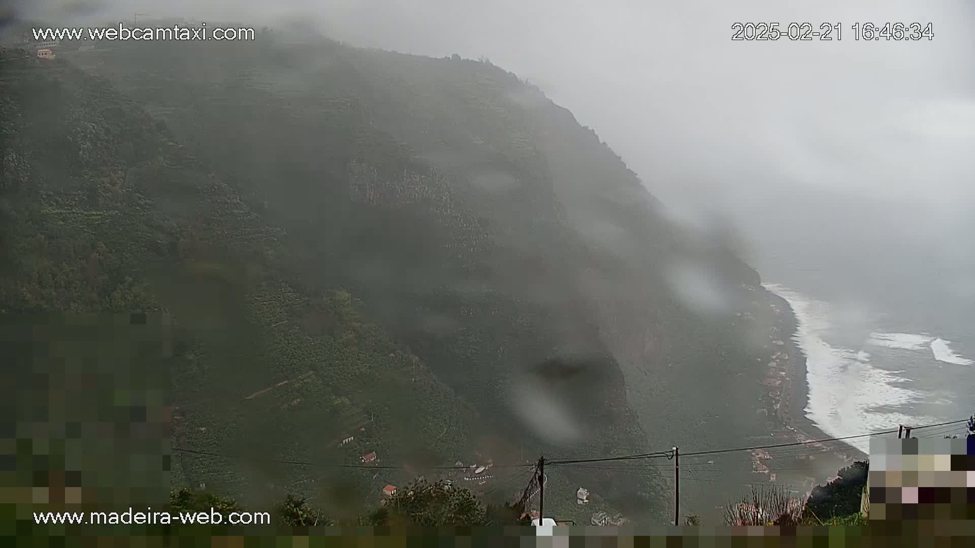 A rain-swept coastal mountainside descends to a rocky shore with crashing waves under a misty sky.