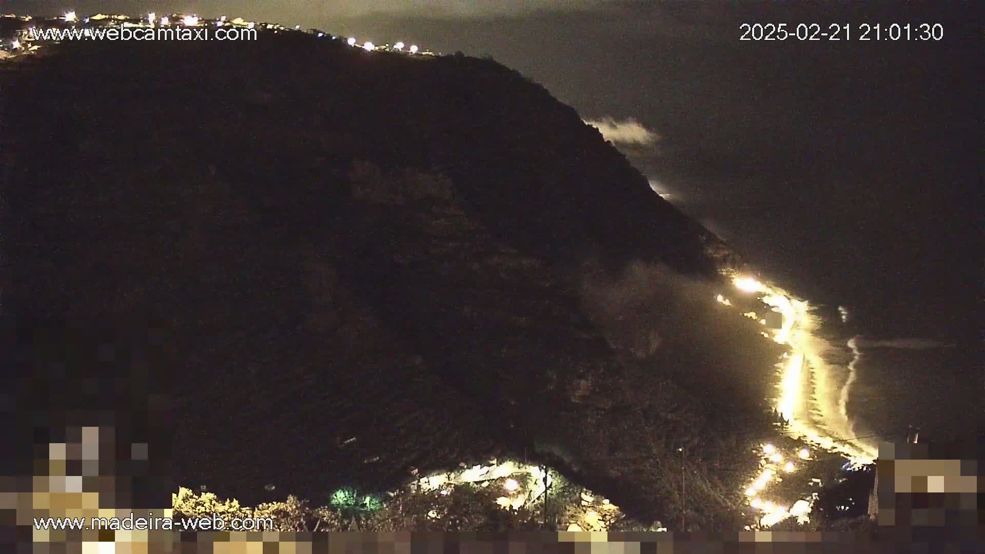 A nighttime, coastal view shows a dark cliffside with a road and some lights along the water's edge, under cloudy conditions.