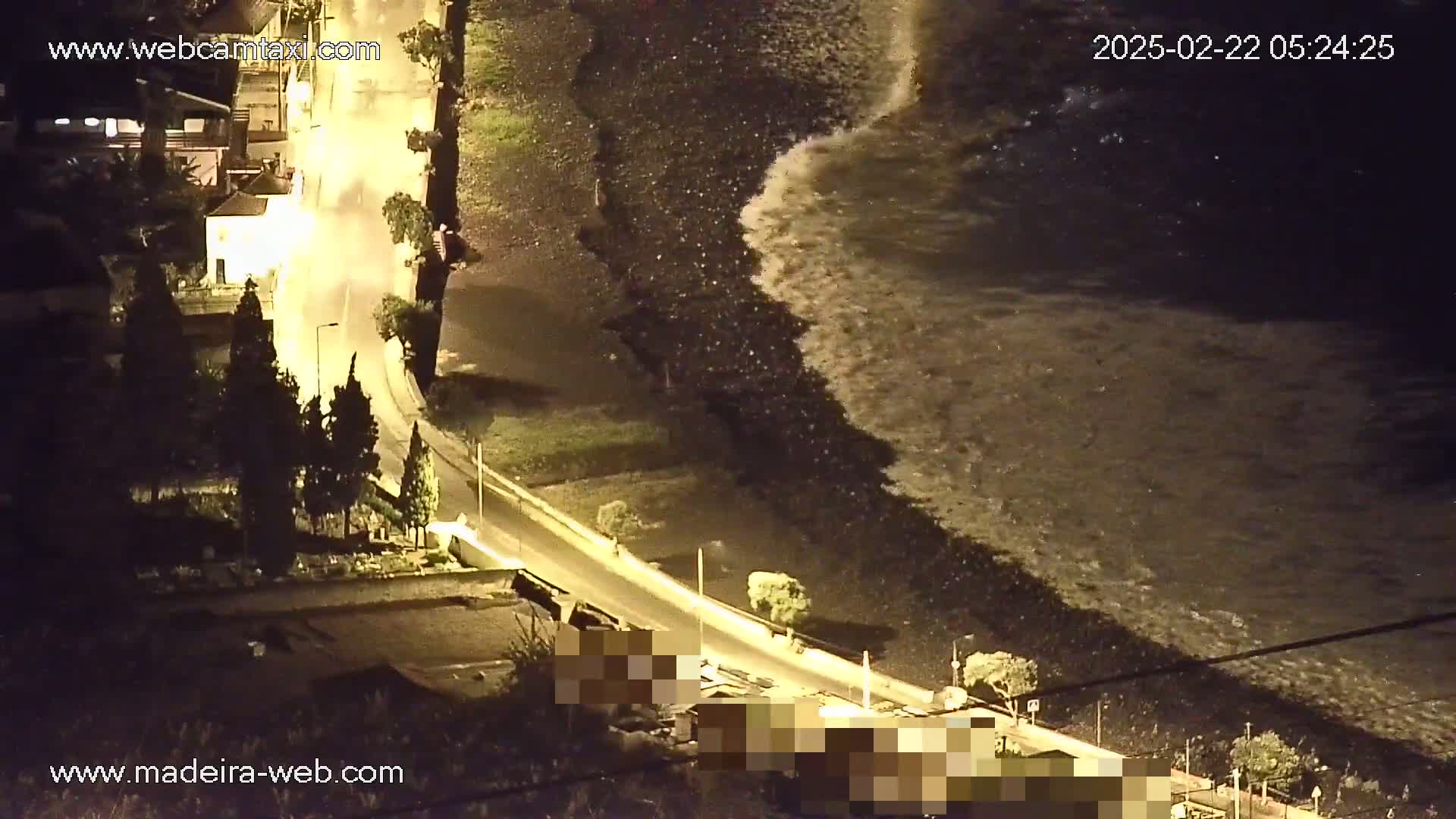 At night, large waves crash on a dark beach next to a road with cars parked alongside it, illuminated by streetlights.