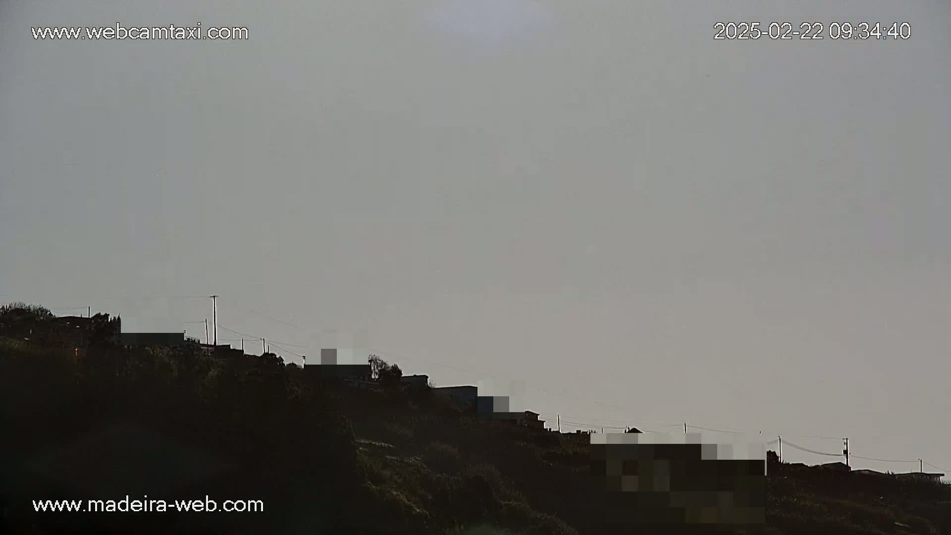 A hillside silhouetted against a pale gray sky shows several buildings and power lines under overcast conditions.