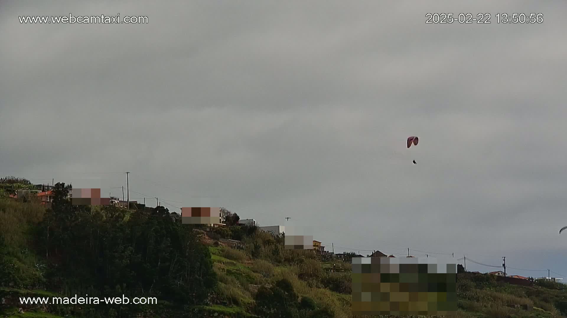 A paraglider flies over a hillside village under an overcast sky.