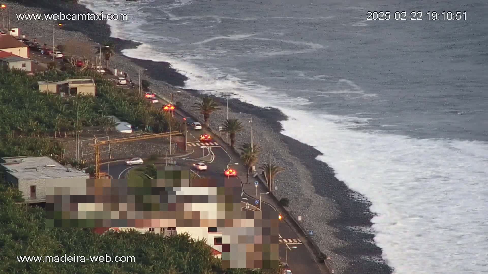 A coastal road with cars and palm trees runs alongside a dark-colored, pebbly beach where waves are breaking against the shore under somewhat overcast conditions.
